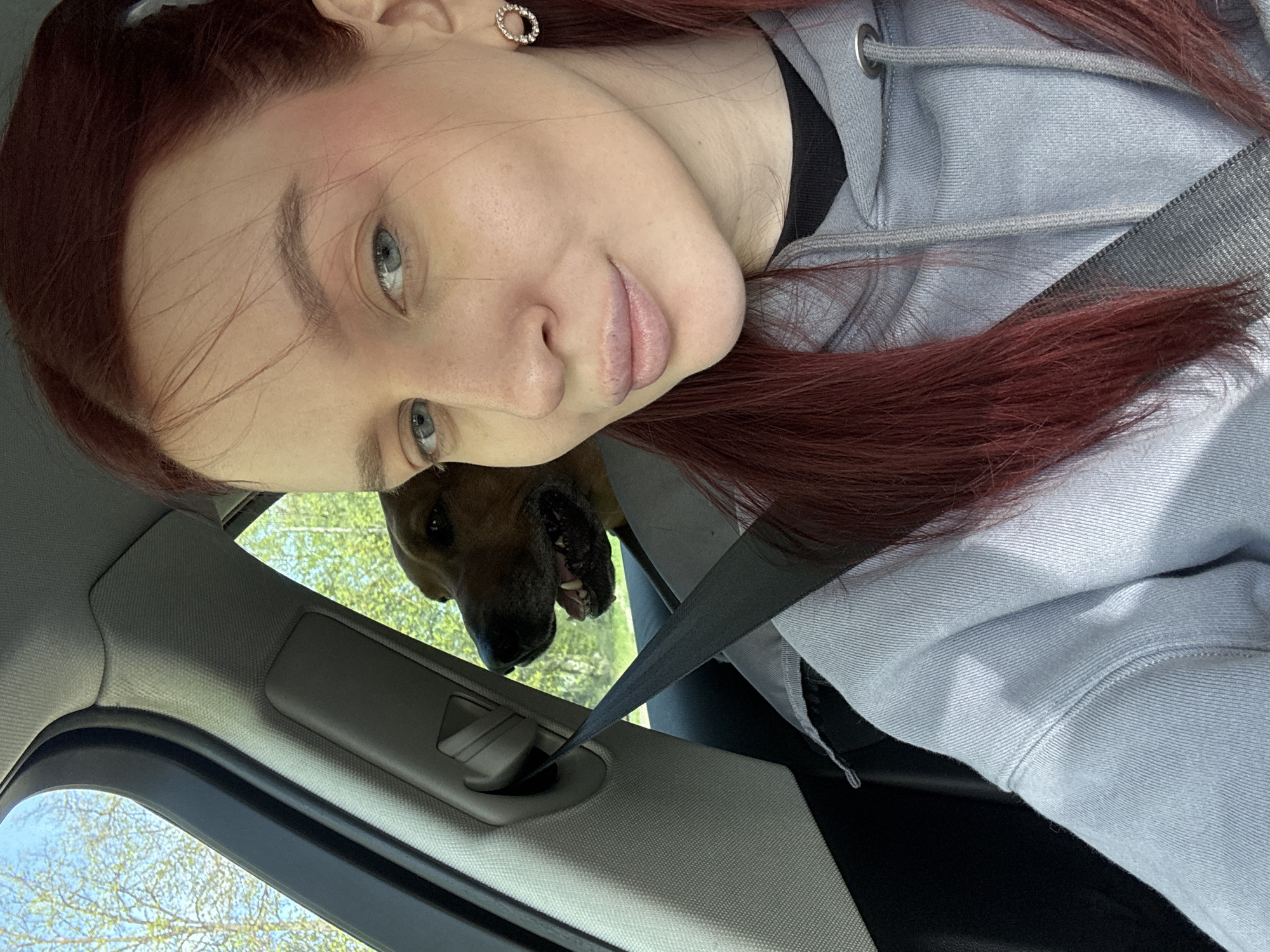 A woman with red hair is in a car, wearing a grey hooded sweatshirt and a seatbelt. A dog is partially visible behind her, peering into the car from outside. The car's interior is visible, including the door and window frame. Outside the car, trees and foliage are visible. The location of the photo is not determinable from the image alone.