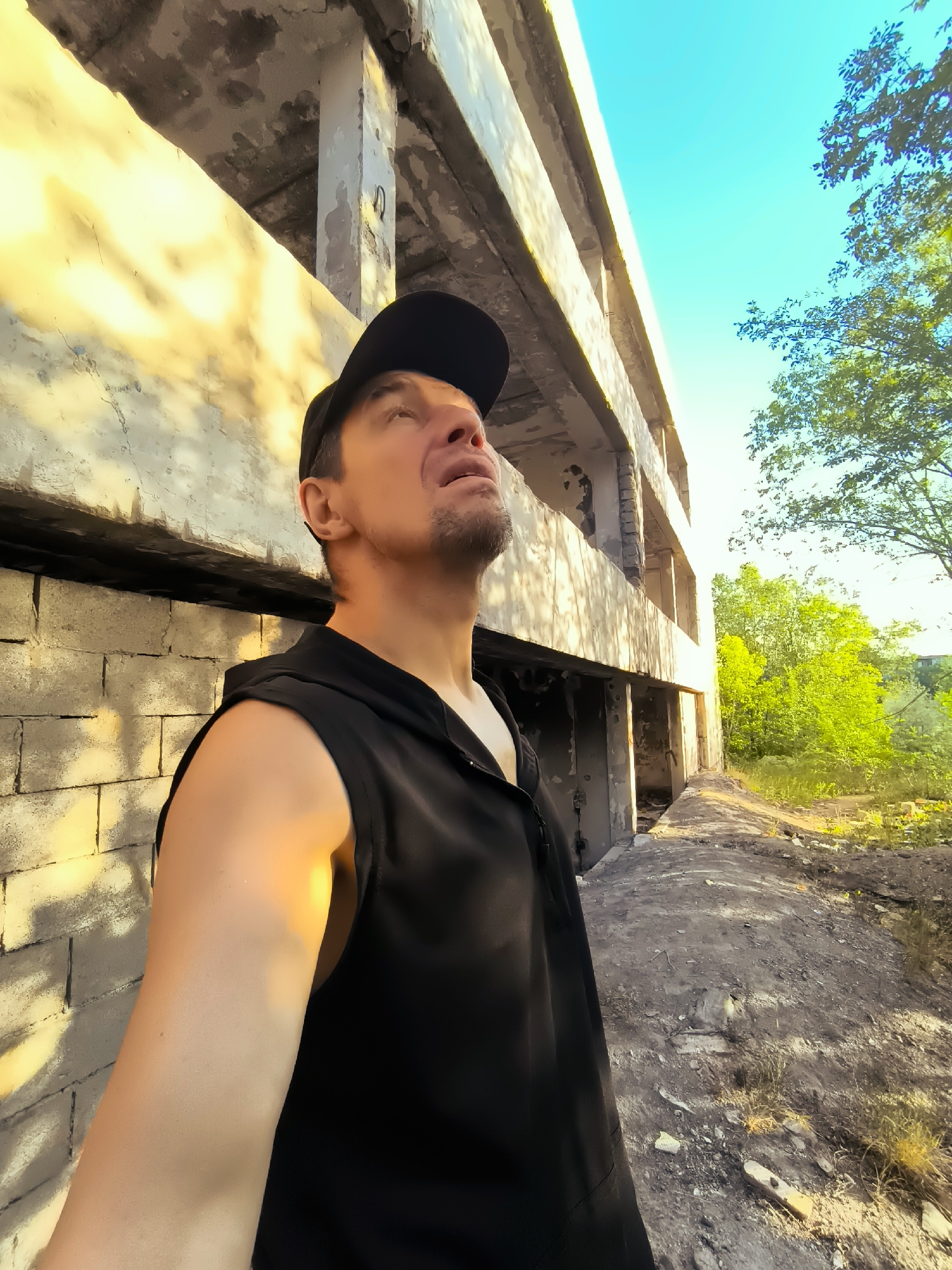 A man wearing a black sleeveless hoodie and a black baseball cap is standing in front of an abandoned concrete building. The man is looking upwards towards the sky. The building appears to be under construction or has been partially demolished, with exposed concrete and rebar visible. The surrounding environment is overgrown with vegetation and debris, suggesting an area that has been neglected or is in disrepair. The lighting in the image indicates it is daytime, with sunlight casting shadows on the building's facade. The location appears to be outdoors, possibly in an industrial or derelict area of Qarağandı city administration, Kazakhstan.