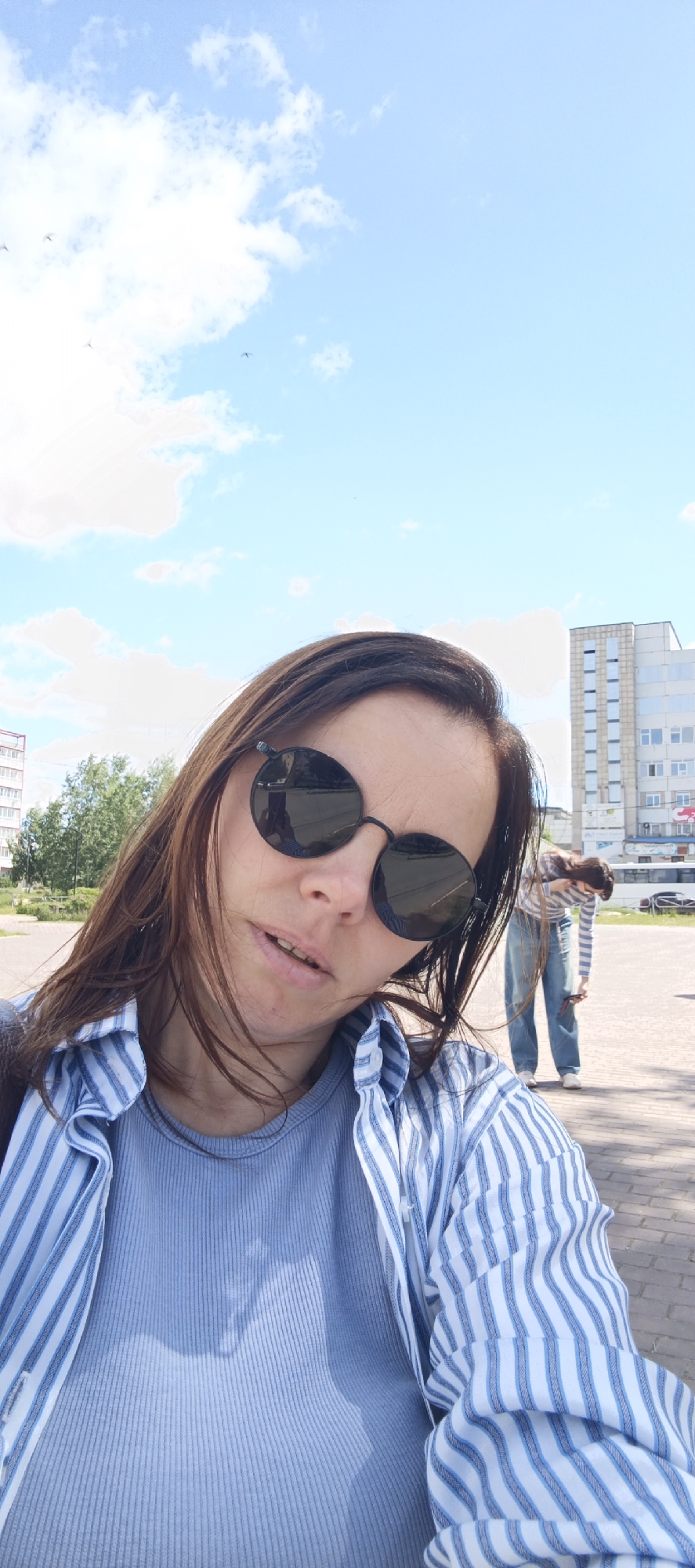 An outdoor selfie captures a woman with long brown hair, wearing round sunglasses, a blue ribbed tank top, and an unbuttoned blue and white striped shirt. Her head is tilted, and her mouth is slightly open. In the background, another person in similar attire is bending over, appearing to pick something up from the ground. Behind them, a tall, modern building with many windows stands under a bright blue sky with scattered white clouds. Trees and a paved area are also visible. The setting is likely a public urban space in Chaikovsky, Russia, possibly a plaza or street, given the architecture and open sky.
