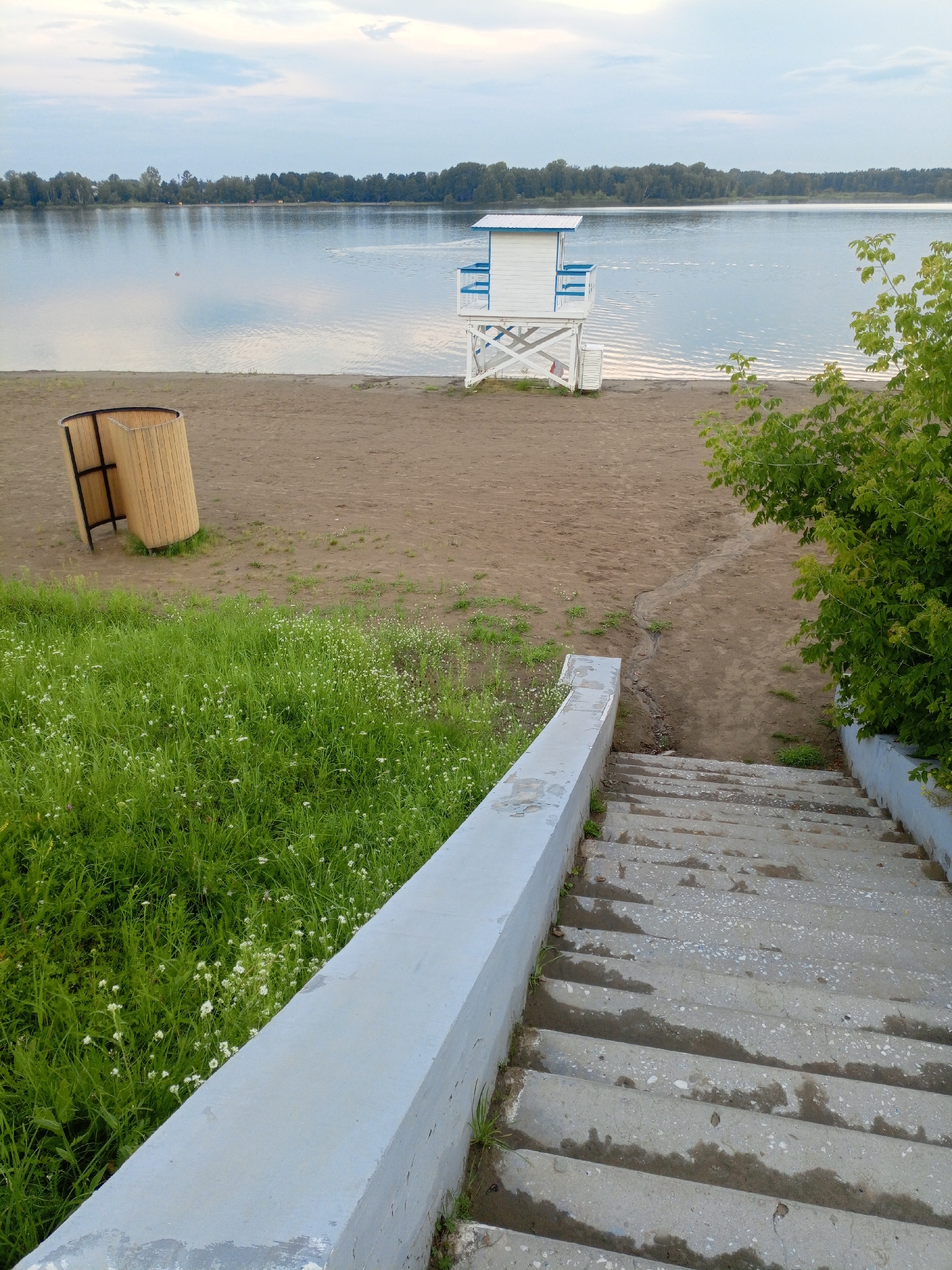 The image depicts a calm lake or wide river scene with a sandy beach and lush green grass. In the foreground, a set of concrete stairs with a weathered light grey railing descends from the viewer's perspective. The stairs show signs of wetness and some minor vegetation growing at the edges.
To the left of the stairs and further up on the beach, there is a curved, light-colored wooden structure, possibly a changing booth or shelter. A small, white and blue lifeguard station stands on the sandy beach near the water's edge. The station has a tall, slender white base with crisscrossing supports and a small white structure with blue trim on top, suggesting it's an observation point.
The water is calm and reflects the cloudy sky and a distant treeline. A few small dark objects, possibly ducks or debris, are visible on the water. A dense line of green trees forms the horizon across the water. To the right of the lifeguard station, a vibrant green bush or tree with dense foliage partially obscures the view of the water and beach. The overall impression is a serene, possibly public, recreational area by the water.
This location is likely part of the beach area along the Angara River in Zheleznogorsk, Russia, known for its scenic natural beauty and recreational opportunities.