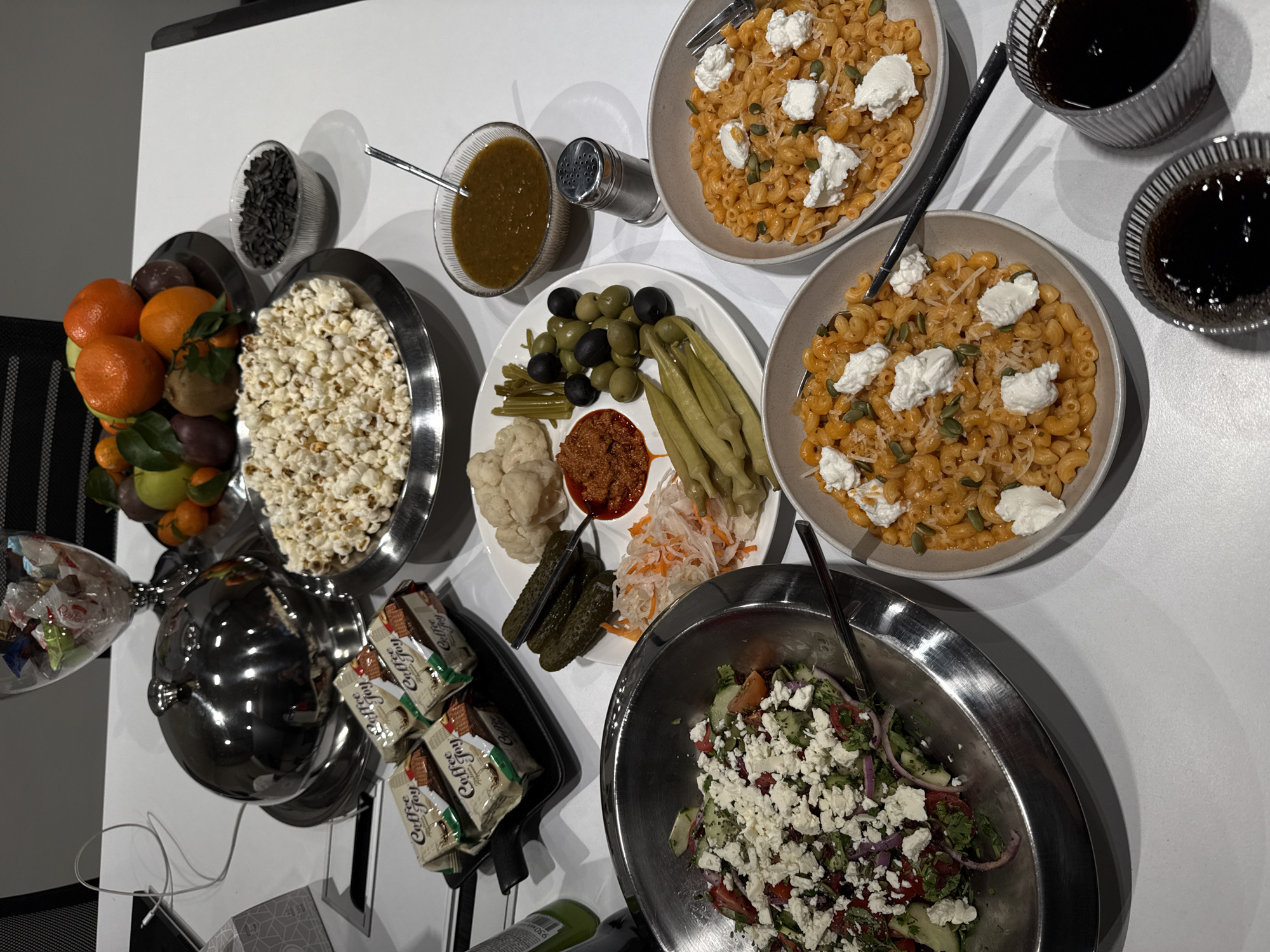 The image displays an indoor tabletop spread of assorted food items in what appears to be an office or meeting room setting in the city of Yerevan, Armenia. A large silver, domed food warmer is centrally positioned on a silver tray. To its left, a black rectangular tray holds several individually wrapped packages of "Coffee Joy" crackers, alongside a coiled white power cable. A clear glass pedestal bowl in the upper left contains various wrapped candies. In the upper right, a silver bowl is filled with fresh fruits, including oranges, apples, kiwi, and mandarins. A small clear glass bowl of black sunflower seeds is adjacent to the fruit. A large silver bowl overflows with white popcorn in the center-right. Below the popcorn, a clear glass bowl with a spoon contains a dark green/brown sauce, accompanied by a silver salt shaker. A large white platter centrally features a selection of pickled items: whole cucumbers, cauliflower florets, green and black olives, a red relish, pickled green beans or okra, and shredded pickled cabbage with carrots. To the lower left, a large silver bowl contains a salad of chopped cucumbers, tomatoes, red onions, and herbs, generously topped with crumbled feta cheese and equipped with a serving spoon. Two beige bowls in the lower-center and right contain macaroni pasta in an orange sauce, garnished with white dollops of cheese and green pumpkin seeds, each with a fork. Two ribbed glass cups containing a dark liquid are located in the lower right. A segment of a dark mesh office chair back is visible in the upper left corner.
