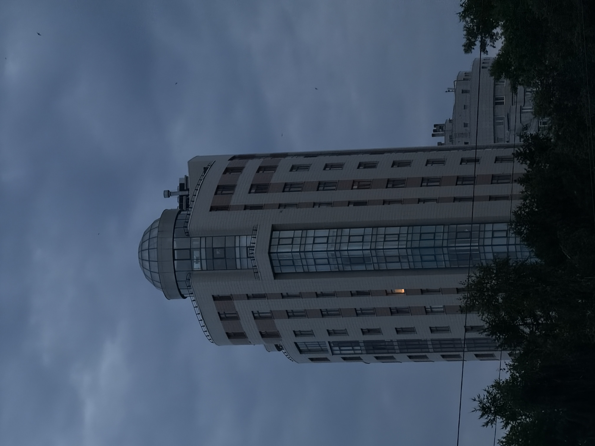 A tall, modern skyscraper with a domed roof stands against a cloudy, dusk sky. The building is light gray with darker accents around the windows and vertical sections. A vertical strip of blue glass windows runs up the center of the main facade. One window on the left side of the building emits a warm, orange light. In the distance, another, shorter, circular building is visible. Silhouetted trees and power lines are in the foreground. Two birds are visible in the sky. The scene is consistent with the residential skyscraper "Riviera" located on the bank of the Iset River in Yekaterinburg, Russia.