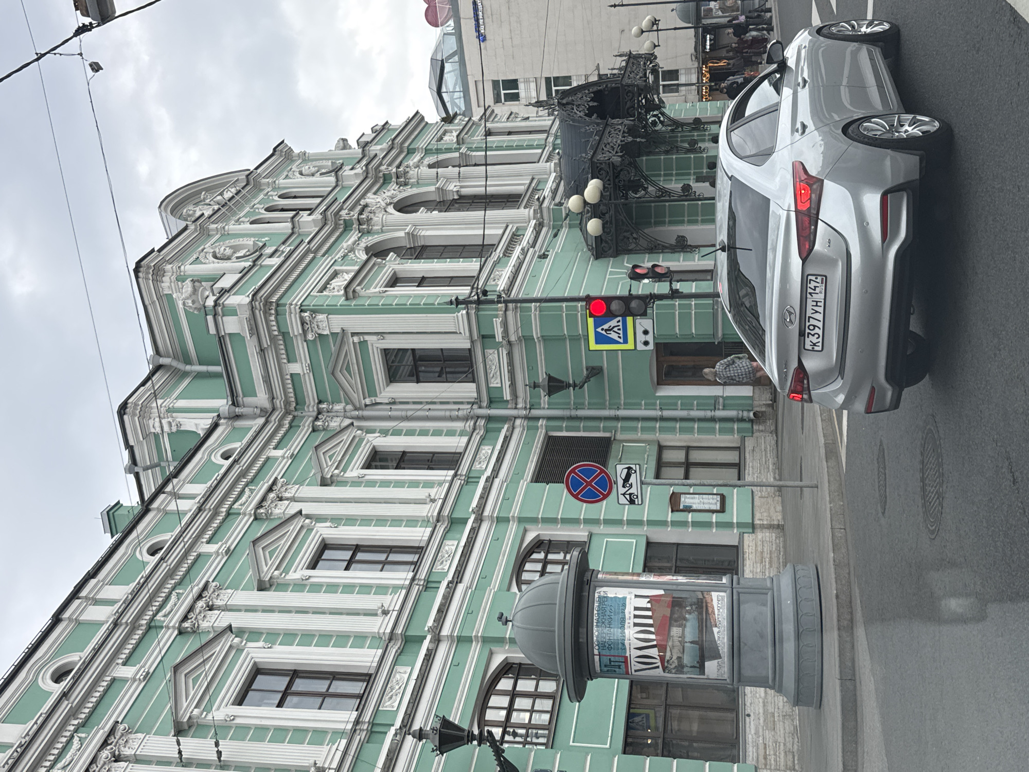 The image captures a street scene in Saint Petersburg, Russia, featuring a prominent, ornate building painted in a pale green hue with white decorative elements. The architecture suggests a historic European style, with multiple windows, columns, and intricate carvings. A silver sedan is visible in the foreground, driving on the wet street, its license plate reading "K397YH 147 RUS". Traffic lights are present, with a red light illuminated. A pedestrian walkway is marked with a crosswalk sign. A large cylindrical advertising pillar stands on the sidewalk, displaying posters. In the background, other buildings and a glimpse of a modern glass-domed structure are visible. The sky is overcast, indicating a cloudy day.