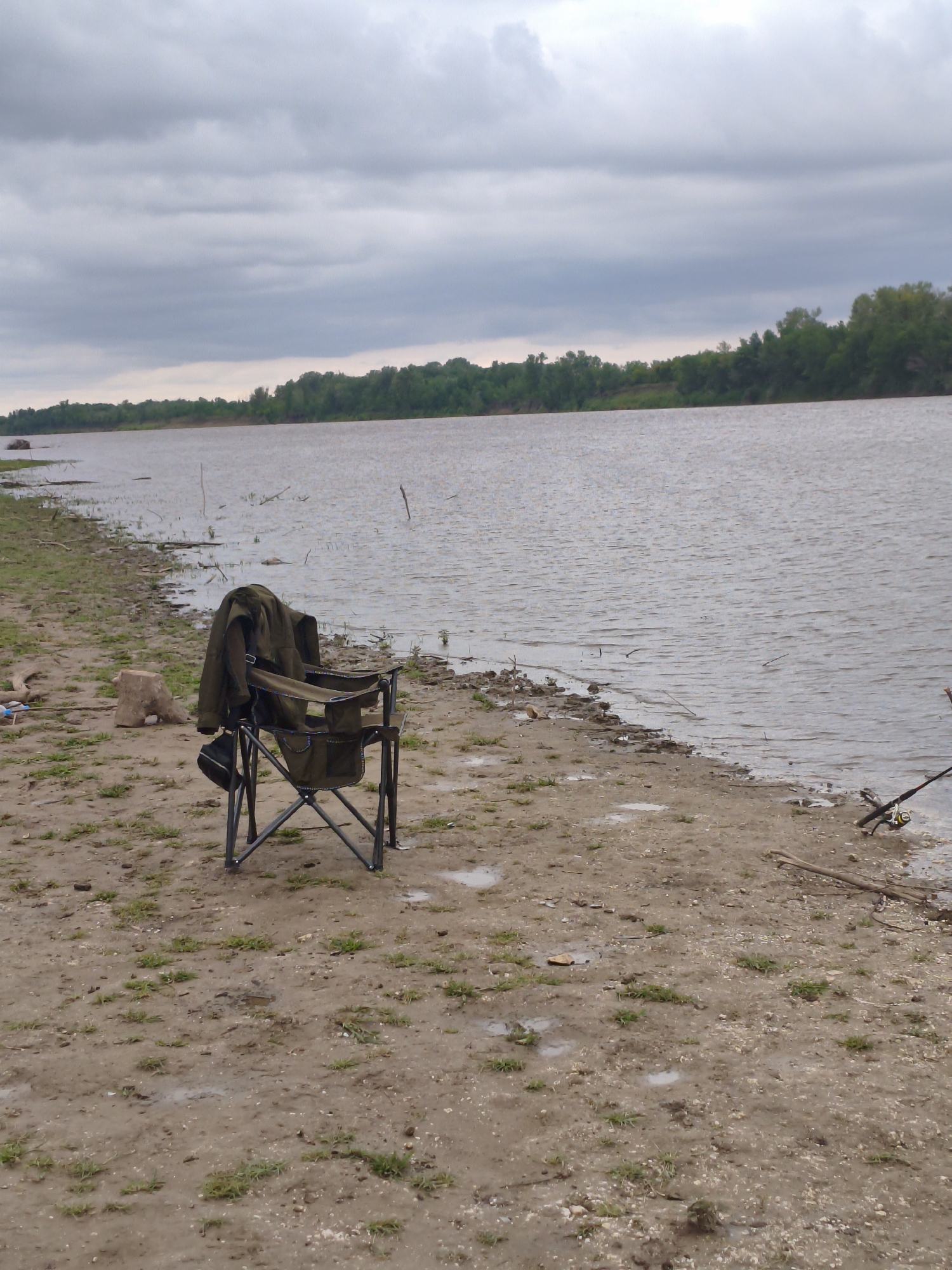 The image depicts a lakeside scene, likely near the village of Aksuat in the Aksuat rural district of Kazakhstan. A dark green fishing chair, folded and with a jacket draped over its back, stands on the muddy, sparsely grassed shore. To the right of the chair, on the edge of the water, are two fishing rods. The water is a murky brown, with small waves. The opposite bank is lined with dense green trees. The sky is overcast with grey clouds, suggesting a cool or rainy day. The overall atmosphere is one of quiet anticipation, suggesting someone is preparing to fish or has recently finished.