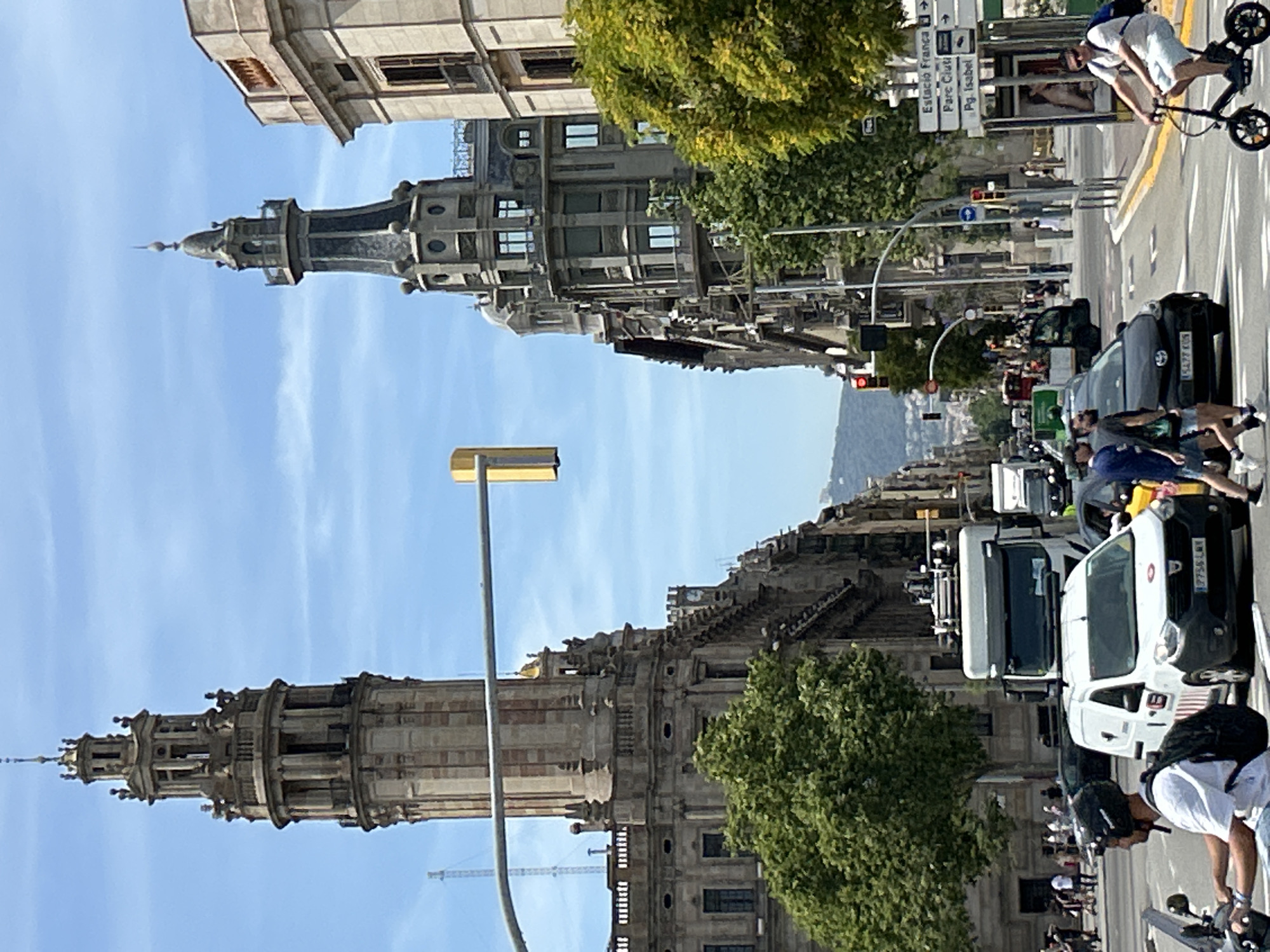 This image captures a street scene in Barcelona, Spain, likely in the Eixample district, given the architectural style and street signs. The photo is taken from street level, looking down a wide avenue.
Dominating the left side of the frame is a tall, ornate building with a prominent tower, characterized by classical architectural elements like columns and decorative stonework. On the right side, a similarly grand building features a distinct spire-like tower with darker, possibly slate, roofing and circular decorative motifs. Both buildings exhibit the characteristic architecture of late 19th and early 20th-century Barcelona.
The street is busy with vehicular traffic. A white truck and a white van are visible, as are several cars, including a black hatchback. A person is riding a black electric scooter in the foreground on the right, wearing sunglasses and light-colored shorts. On the left, a person on a motorcycle, wearing a helmet and backpack, is in the process of turning. Pedestrians are walking on the sidewalks.
Traffic lights and street signs are present. One signpost indicates directions to "Estació França," "Parc Ciut," and "Pg. Isabel," which are landmarks in Barcelona. Lush green trees line the street, providing a contrast to the urban architecture. The sky is bright blue with scattered white clouds, suggesting a sunny day. In the distance, a hazy mountain range is visible. The overall impression is of a vibrant, historic urban environment.