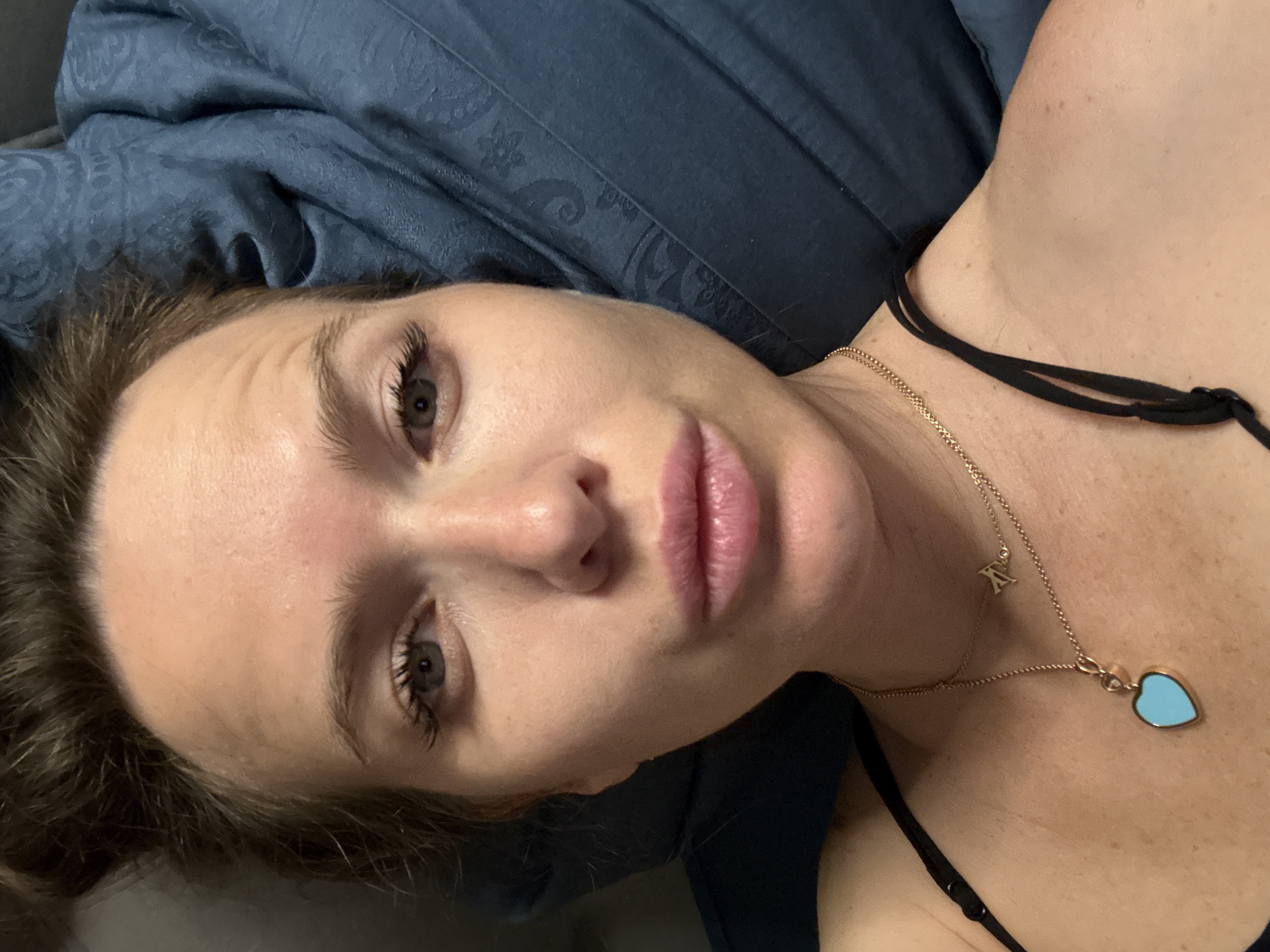 A close-up selfie shows a woman lying on a blue patterned bedspread. She has light skin, brown hair, and pink lips. She is wearing a thin black tank top and two gold necklaces, one with a heart-shaped pendant. The location of the image is unspecified in the prompt, and therefore the reference to the city of Khimki, Russia is extraneous and irrelevant.