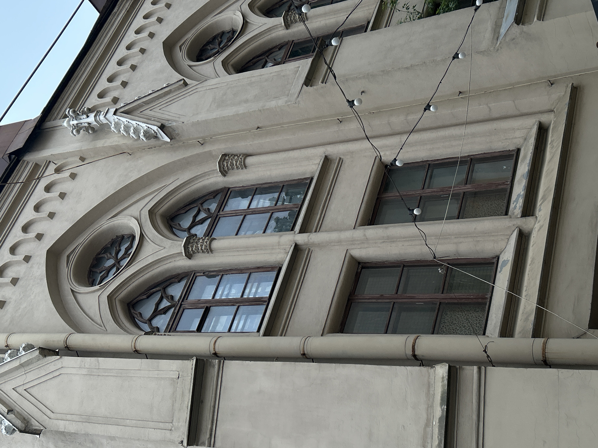 The image depicts a section of the architectural facade of a building in Saint Petersburg, Russia, showcasing elements of neo-Gothic design. The facade is primarily rendered in a light grey hue. The building features decorative arched windows with intricate mullions. A string of lights is draped across the facade, with visible bulbs and wiring. Architectural details such as decorative moldings, triangular pediments, and a gargoyle adorn the building. A horizontal pipe runs across the lower portion of the image. The photograph is taken from a low angle, emphasizing the height and details of the building.