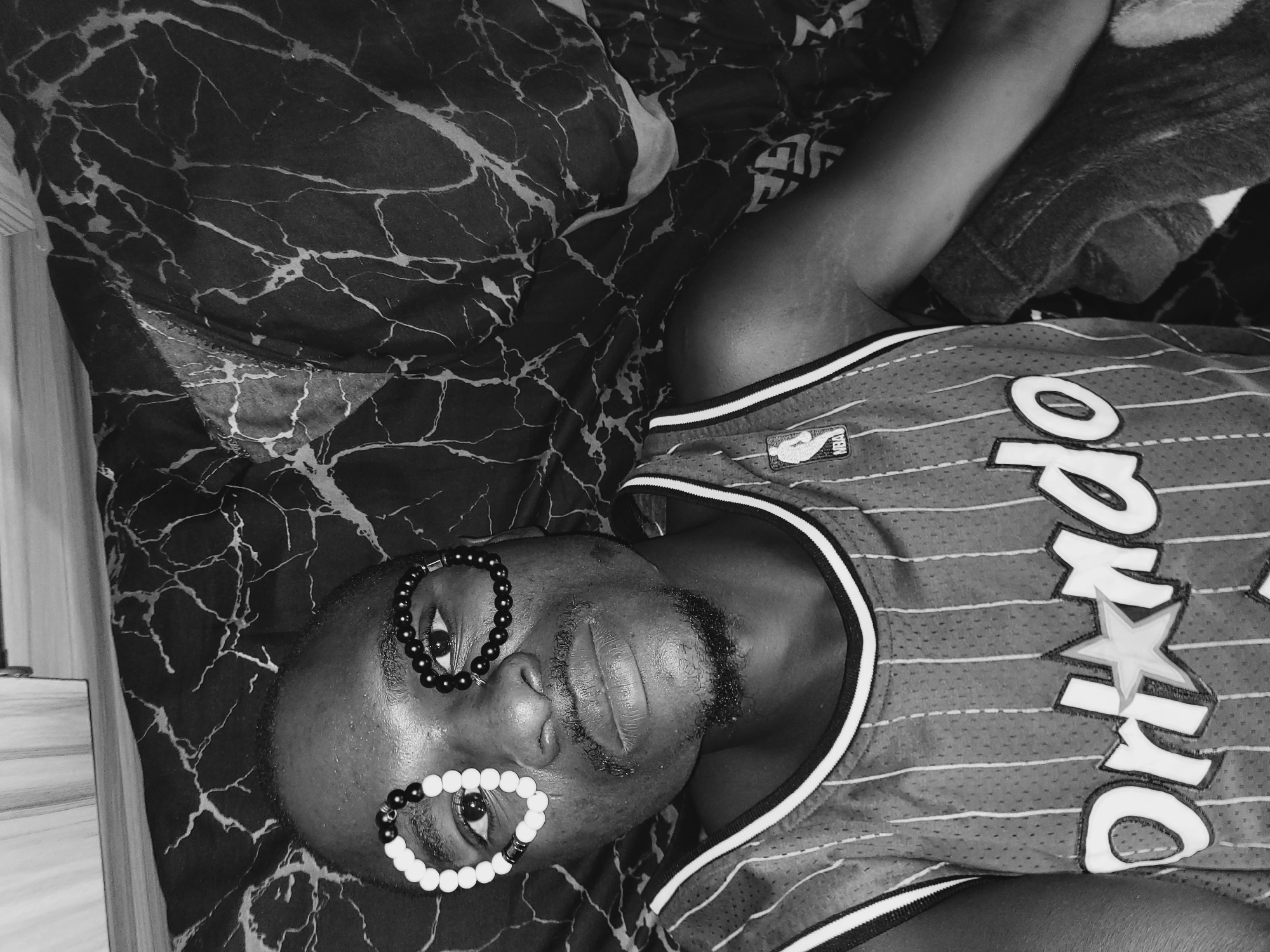 A black and white, high-angle, eye-level shot captures a person lying on their back on a marbled patterned surface. The person is wearing a striped basketball jersey and has bracelets placed around their eyes. The person has a goatee and appears to be looking upwards. The scene is set indoors, possibly in Bukuru, Nigeria, though no specific city landmarks are visible. The image has a grainy texture.