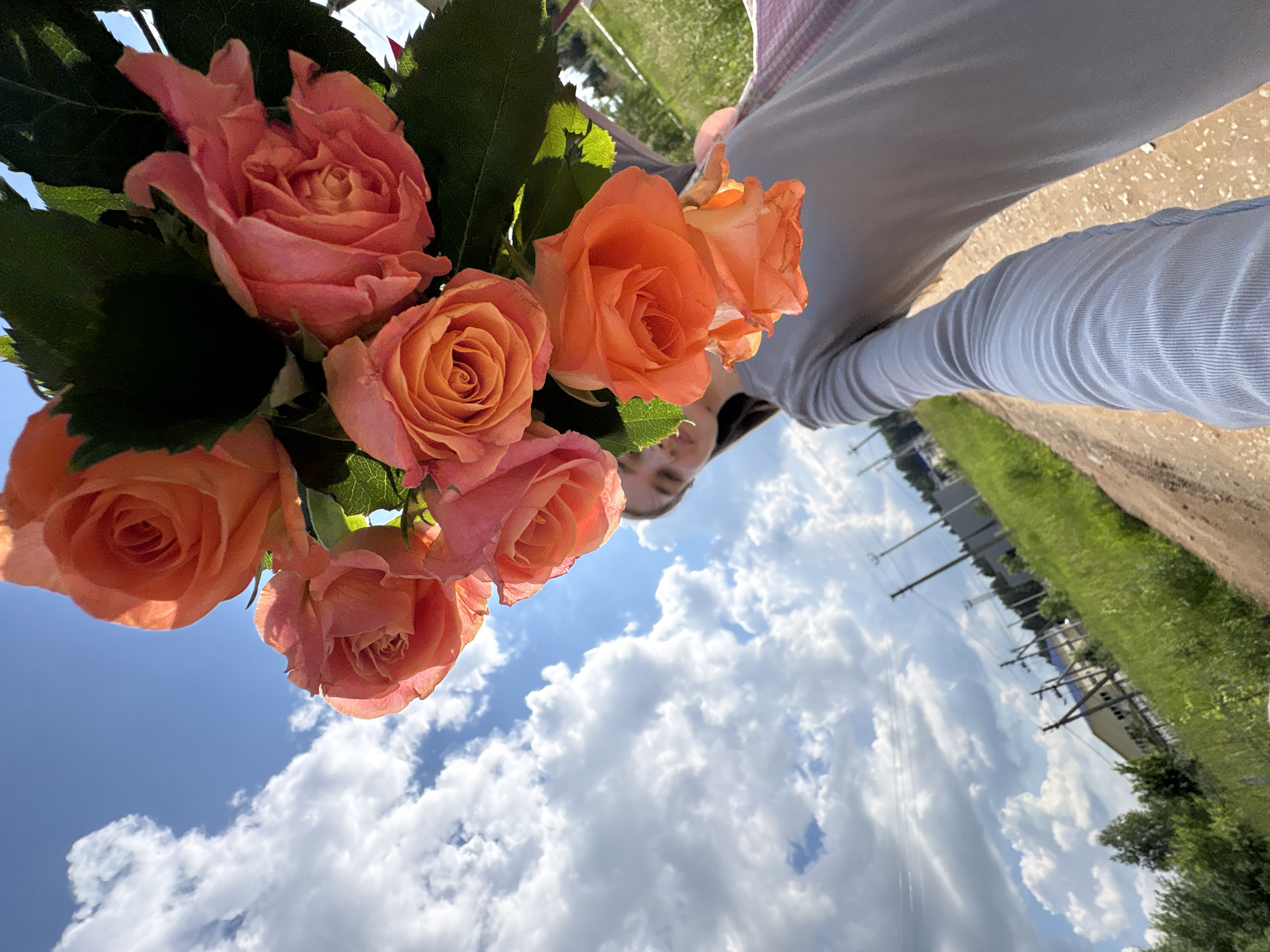The image shows a first-person perspective of a person holding a bouquet of orange and pink roses against a blue sky with white clouds. The person is wearing a gray long-sleeved shirt and has a pink garment draped over their shoulder. The bouquet is in the foreground, obscuring the person's face and upper body. In the background, a dirt road leads into a grassy area with trees and some buildings, suggesting a rural or semi-urban setting. Electrical poles and wires are visible in the distance. The scene is outdoors during daylight.