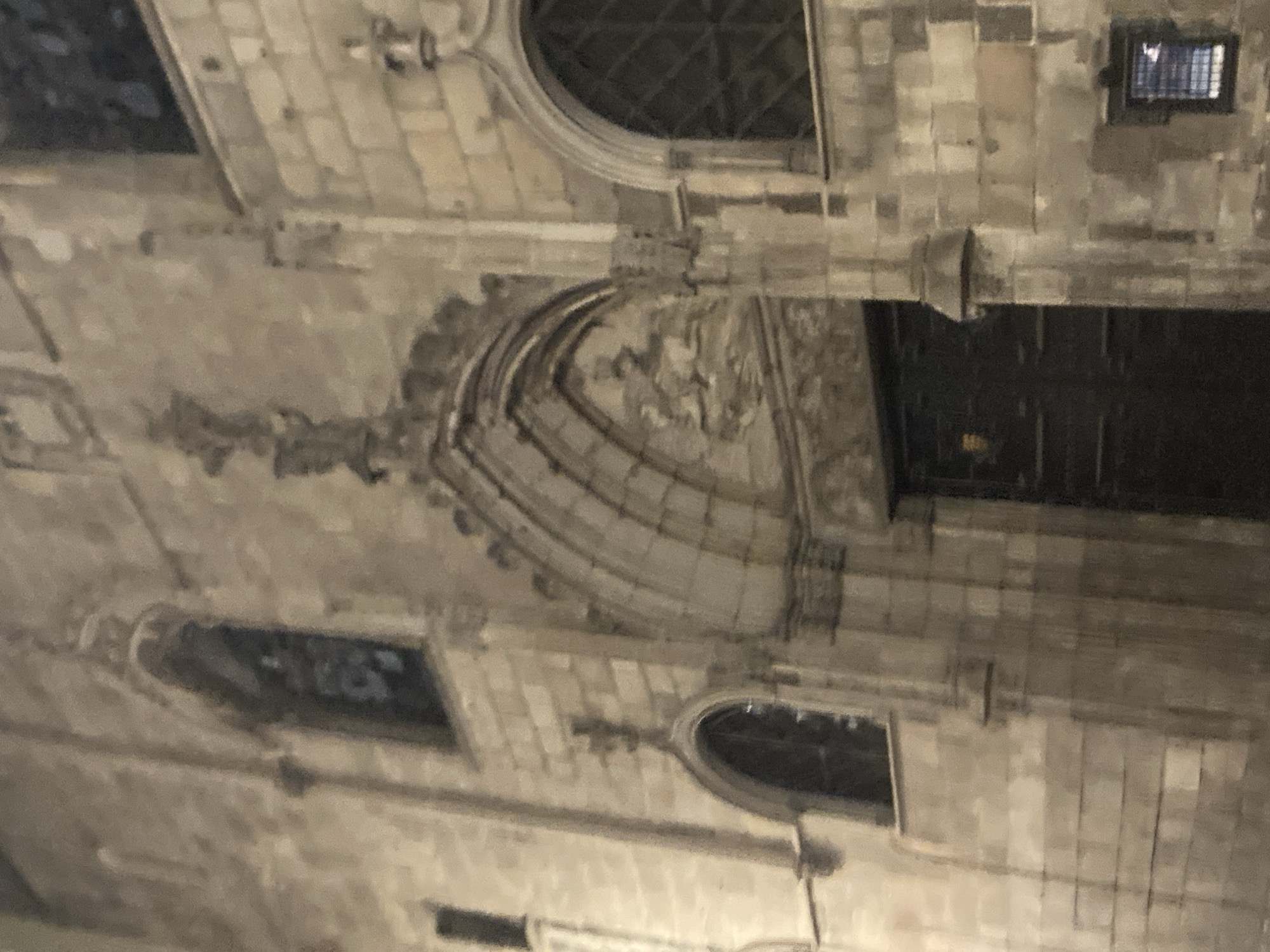 The image shows the exterior of a stone building, likely a church or a historic landmark, at night. The architecture features Gothic arches, ornamental carvings, and windows with decorative grilles. The building's facade is constructed from large, weathered stone blocks, giving it an aged appearance.
The main entrance is a large, dark wooden door set within a prominent arched portal. Above the doorway, there are intricate stone carvings, possibly depicting religious or historical scenes. Several arched windows are visible on the facade, some of which are dark and appear to be stained glass. Small, square windows with gratings are also present.
The lighting in the image is dim, suggesting it was taken at night or in low-light conditions. The overall tone of the image is muted, with shades of gray and beige dominating the color palette. The stone appears damp, possibly due to rain or dew, reflecting the ambient light.
Based on the architectural style, this building is likely located in Barcelona, Spain. The Gothic architecture and ornate stonework are characteristic of many historic structures in the city. Without more specific information or visual cues that definitively link it to a particular landmark, it is challenging to pinpoint the exact location. However, it resembles elements found in structures such as the Barcelona Cathedral or other medieval churches in the Gothic Quarter.