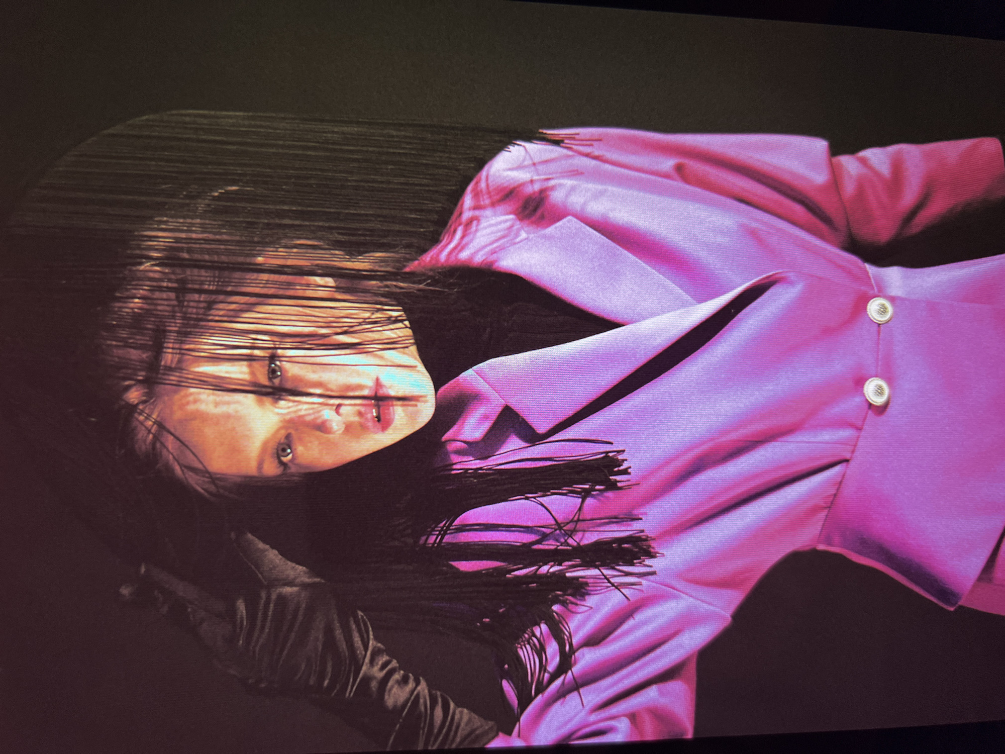 A close-up, head-and-shoulders studio portrait depicts a person with fair skin, styled with dark, fringe-like bangs and a black turtleneck. The subject is wearing a bright pink, satin blazer with two prominent gold buttons. On the left side of the image, a black satin glove is visible, with the subject's left hand emerging from it and resting near their head, holding their hair. The lighting is dramatic, with strong highlights and shadows, creating an artistic and somewhat moody effect. The background is dark and appears to be a solid color, possibly black or a very deep shade of brown, with a subtle curved shadow element on the right. The image appears to be a photograph taken against a backdrop in a controlled studio environment. No specific location in the city of Unknown, Russia is identifiable.