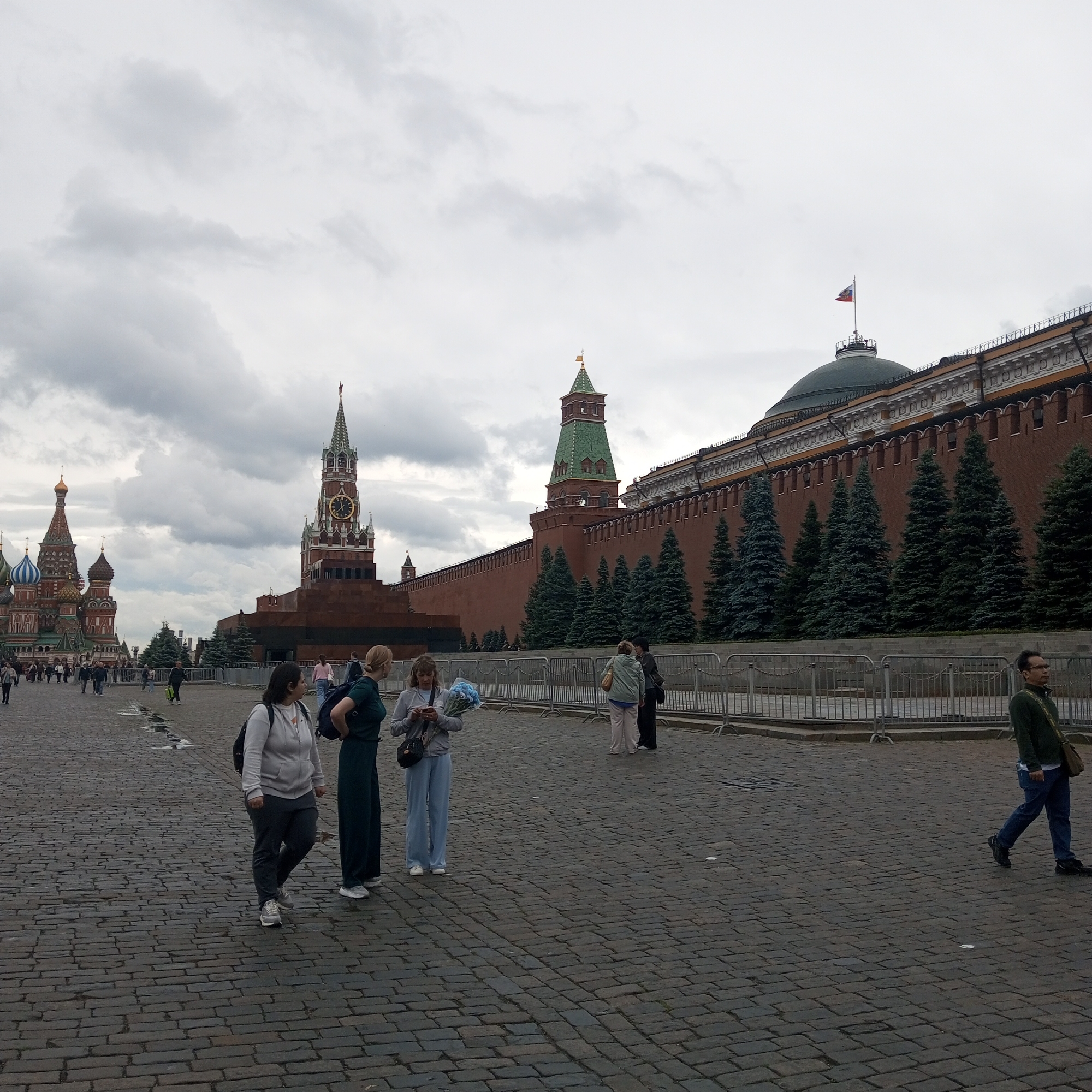 The image shows a cloudy day in Red Square, Moscow, Russia. In the foreground, several people are walking on the cobblestone ground. To the left, St. Basil's Cathedral is visible with its colorful onion domes. In the center, the State Historical Museum and the Lenin Mausoleum are prominent structures. The Kremlin wall extends across the background, with trees planted along its base. A Russian flag flies atop a dome on the Kremlin. The scene is a public space with people engaging in various activities, indicating a tourist or public gathering area.