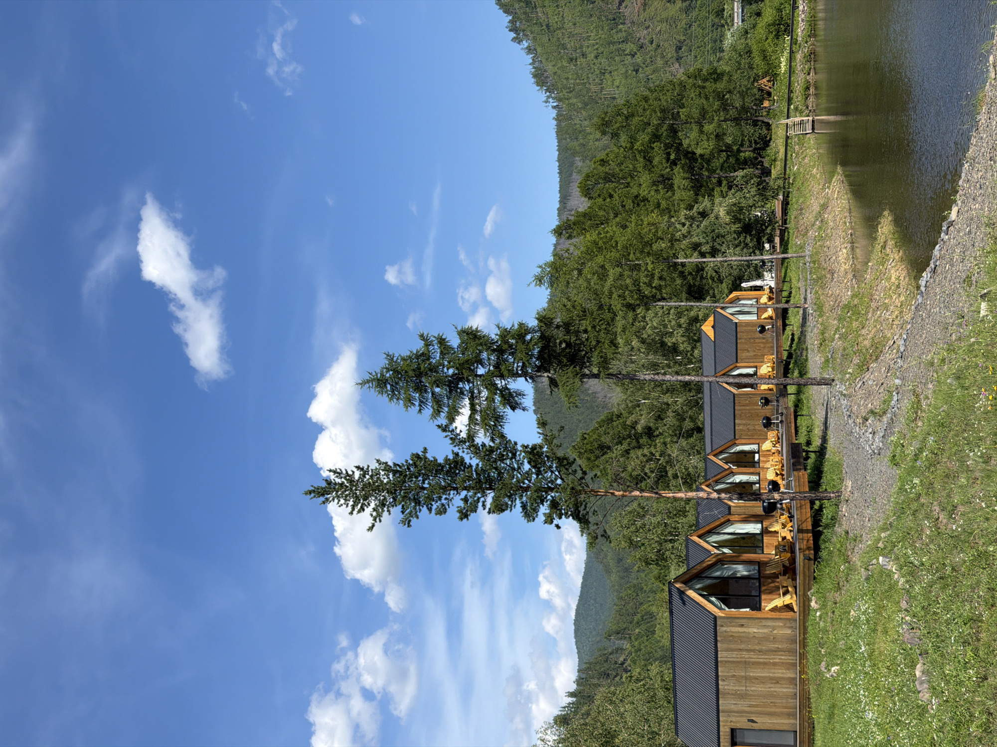 The image depicts a row of modern wooden cabins with black pitched roofs situated on a wooden deck beside a tranquil body of water. The cabins feature large, angled windows offering views of the surrounding natural landscape. Several Adirondack chairs and small grills are placed on the decks, suggesting a recreational setting. Tall evergreen trees flank the cabins, and the background is dominated by lush, green forested hills under a bright blue sky with scattered white clouds. The scene appears to be a resort or glamping site located in the scenic environment of Bol'shoe Goloustnoe, Russia, likely on the shore of Lake Baikal or a nearby body of water.