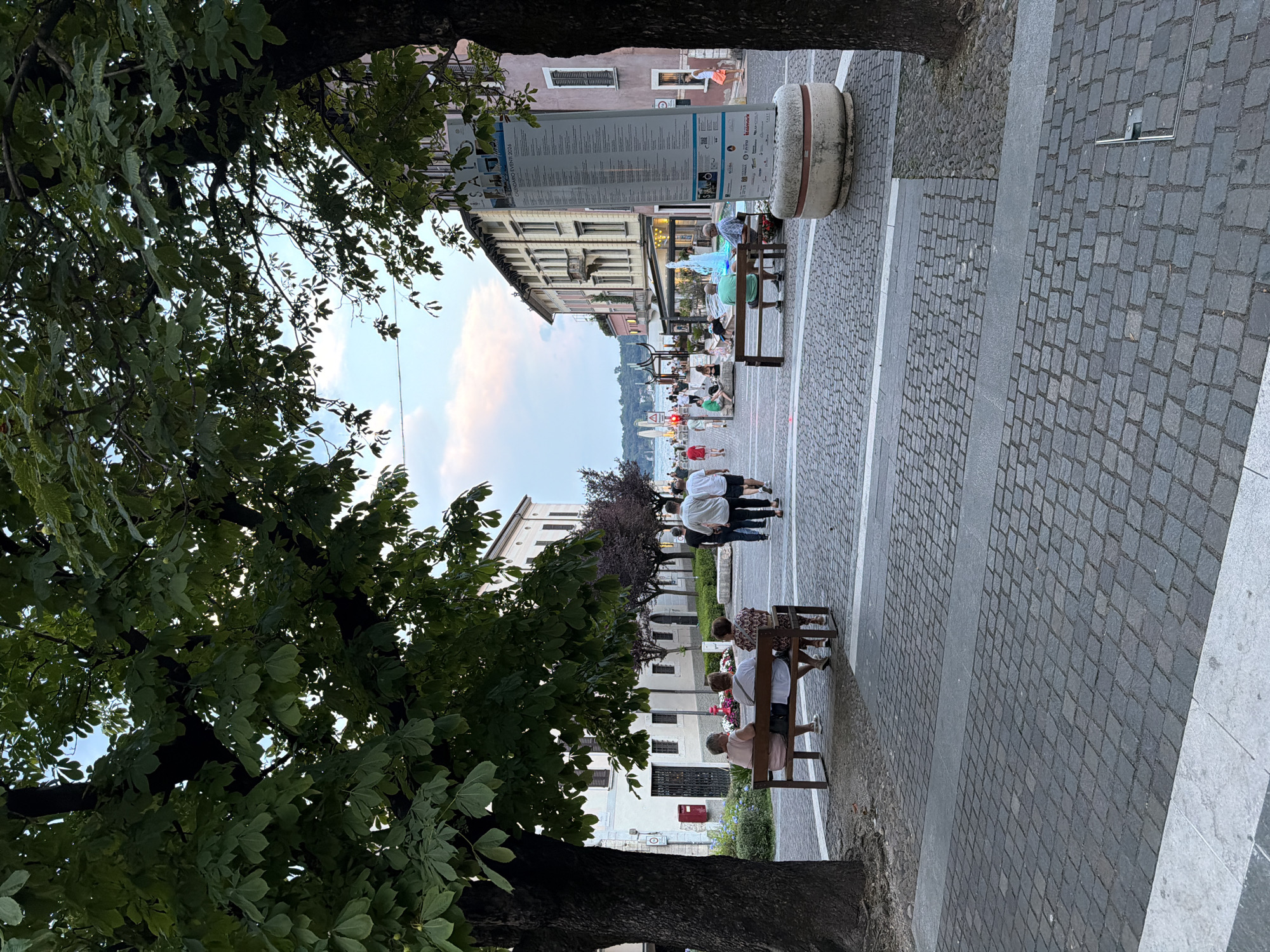 This image depicts a public walkway in Salò, Italy, likely a lakeside promenade given the presence of water and boats in the background. The scene is framed by mature trees with dense green foliage. A cobblestone path forms the foreground and middle ground, transitioning to a lighter stone surface in the immediate foreground.

In the middle ground, several people are visible. On the left, three women sit on a wooden bench facing away from the camera, looking towards a building. To their right, a group of people walks away from the camera. Further to the right, a fountain with vibrant blue water is a focal point, with more people seated on benches nearby. A large information board, possibly displaying an event calendar, stands beside the fountain.

The background features buildings lining the street, with a mix of architectural styles, some with balconies and awnings. The sky above is a pale blue with hints of pink and white clouds, suggesting either early morning or late evening. The overall atmosphere is relaxed and leisurely, with people enjoying the outdoor space.