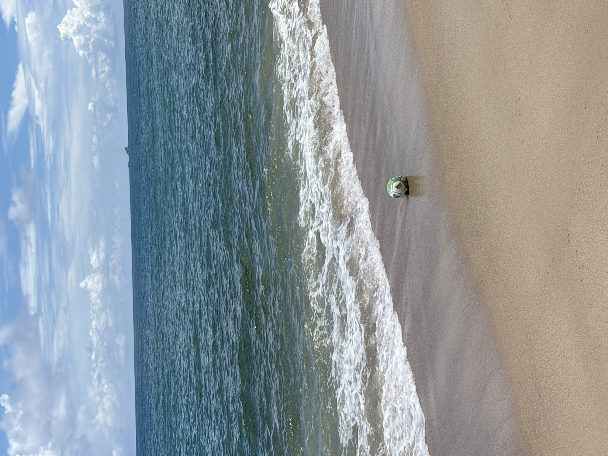 A soccer ball rests on a sandy beach near the waterline. The ocean, a deep blue-green, stretches to the horizon under a sky filled with white clouds. The beach itself is a light tan, with the wet sand reflecting the light. The ball is green and white, with a visible seam pattern. In the distance, a small boat is visible on the water. The scene appears to be a sunny day.