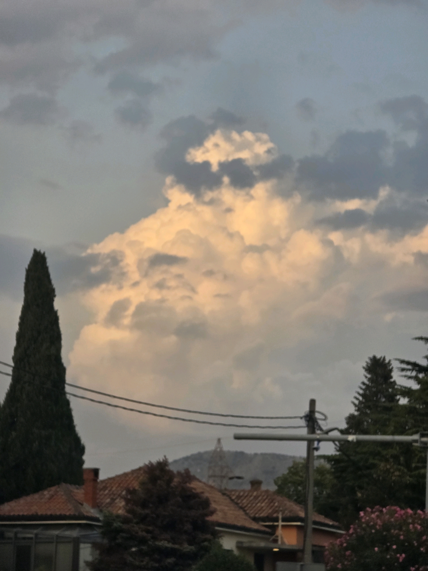 The image depicts a cloudy sky over a cityscape, likely taken at dusk or dawn due to the warm lighting on the clouds. In the foreground, there are terracotta-tiled rooftops and a tall, dark green evergreen tree. A power line stretches horizontally across the middle of the frame. In the background, faint outlines of buildings and possibly a distant structure with a dome or spire are visible, set against rolling hills. The sky is filled with a mix of grey and light orange clouds, with a large, brightly lit cumulus cloud dominating the center. The scene appears to be from Podgorica, Montenegro, given the architectural style of the rooftops and the presence of the tall cypress-like tree, common in the region. There are no people or discernible actions in the image.