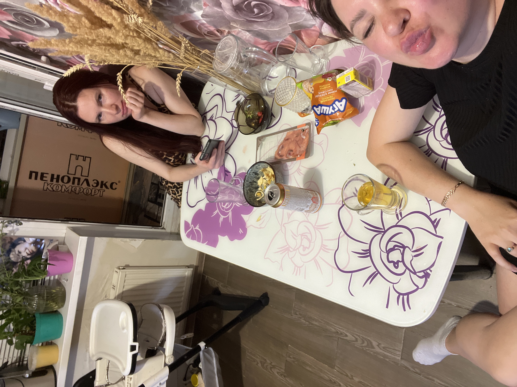 Two young women are gathered around a white table with purple floral decorations. The table is covered in various items, including glasses, a can of what appears to be beer, and a package of snacks. One woman, with long dark hair, is sitting on the left, holding a phone in her hand. The other woman, on the right, is leaning towards the camera with her lips puckered in a playful gesture. She is wearing a black t-shirt and a bracelet. A baby's high chair is visible in the foreground, suggesting a home environment. The scene appears to be indoors, possibly in a dining area or living room. The lighting suggests it could be daytime, but it's difficult to determine the exact time or weather conditions. The overall mood seems casual and relaxed, with a hint of playfulness. The Russian text "ПЕНОПЛЭКС КОМФОРТ" is visible on a cardboard box in the background, which is a brand of thermal insulation materials. The snack package has Russian text that reads "Ариуа" and mentions "мини хрустящие".