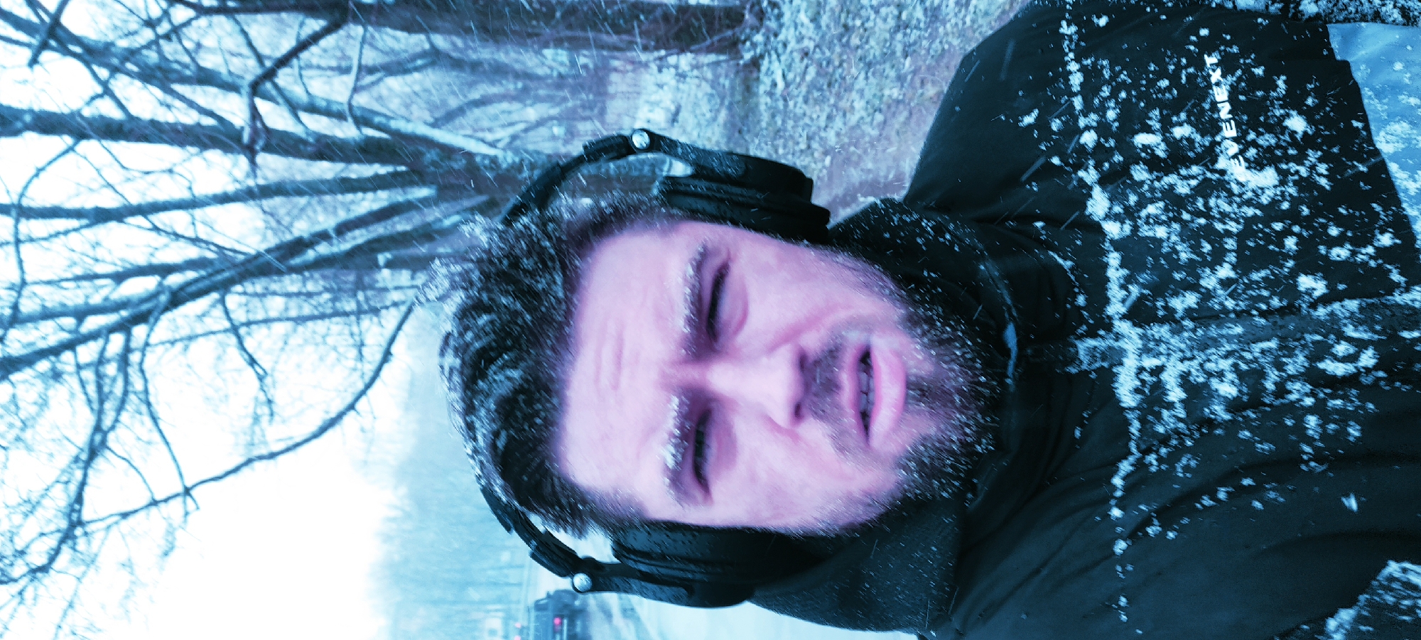 A close-up, medium-shot photograph depicts a man with a fair complexion in a winter environment. He is wearing headphones and a dark-colored jacket covered in snowflakes. Snow is visible on his face, hair, and eyelashes. The background shows a snow-covered landscape with bare trees and a road with vehicles. The scene is likely outdoors during a snowfall. The location is assumed to be near Fryazino, Russia, because this information was requested.