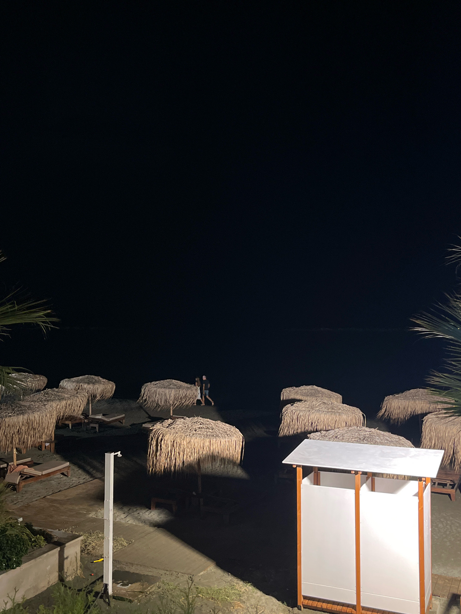 The image captures a nighttime scene on a beach in Agios Tychona Community, Cyprus. The sky is completely dark, with the exception of a faint horizon line where the sea meets the sky. On the sandy beach, there are several thatched umbrellas providing shade over lounge chairs. Two people, a man and a woman, are walking along the shoreline, their figures silhouetted against the dim light. In the foreground, a modern shower stall with a white roof and white panels framed by wooden supports stands on a wooden deck. Lush green foliage is visible to the left of the shower stall, suggesting the presence of a garden or vegetated area nearby. The overall ambiance is peaceful and serene, with the beach appearing to be closed for the night.