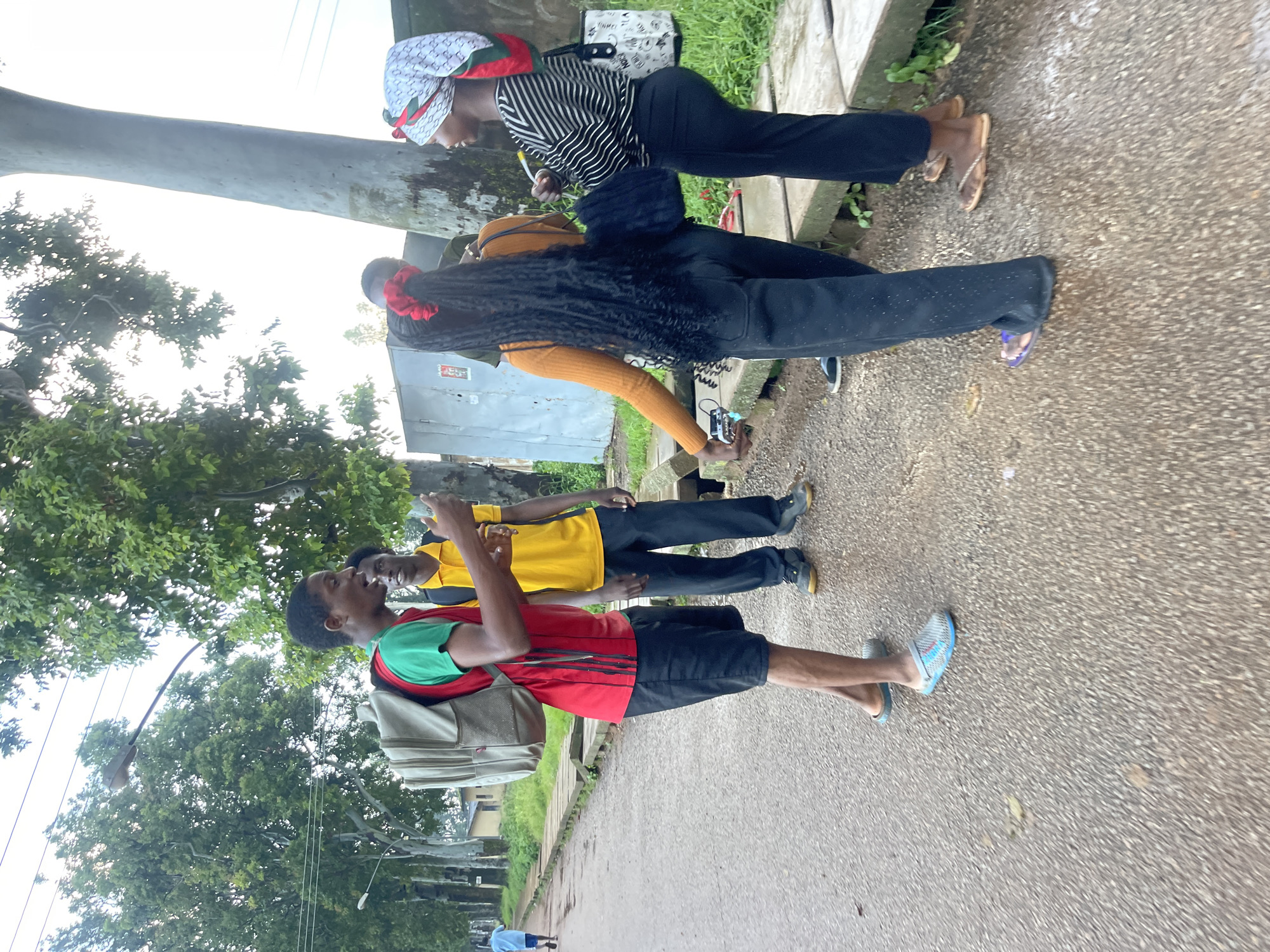 Four young people, two male and two female, are standing on a wet, asphalt road lined with concrete drainage channels and lush green vegetation. A large tree trunk is visible behind the individuals on the right. One male, wearing a red shirt, black shorts, and a backpack, is facing left, gesturing with his hands as if speaking. To his right, another male in a yellow shirt and black pants appears to be listening. Two females are on the right side of the frame. One, wearing an orange long-sleeved shirt and black pants, is facing away from the camera, holding a small device. The other female, in a black and white striped shirt and black pants, is facing left, holding a dark bag. The scene appears to be outdoors, possibly in a residential or semi-urban area, with overhead power lines and street lights visible in the background. The location is identified as Rafin Par, Nigeria.