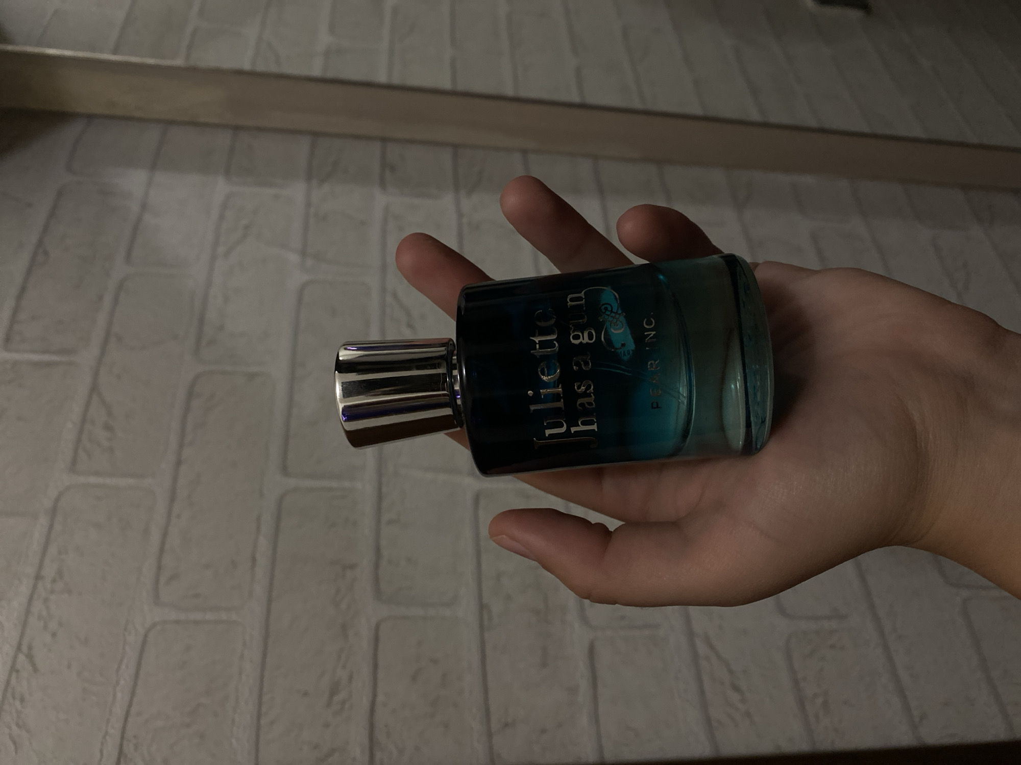 A person's hand holds a glass perfume bottle against a brick-patterned wall. The bottle has a silver cap and a dark blue to teal gradient glass. Text on the bottle reads "Juliette has a gun" and "PEARINC". The background appears to be an indoor setting.