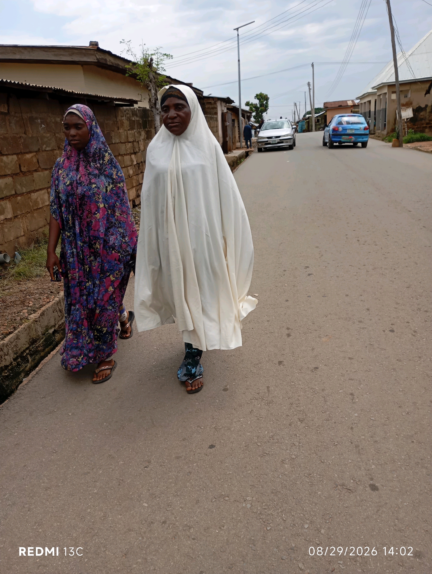 In Shendam, Nigeria, two individuals are walking down a paved road. The person on the left wears a colorful patterned dress and a headscarf, while the person on the right is covered in a white garment and headscarf. The road is lined with buildings on both sides, and there are cars parked and driving in the background. The sky is cloudy, suggesting an overcast day. A watermark at the bottom left indicates the image was taken with a REDMI 13C, and a timestamp in the bottom right shows the date as 08/29/2026 and time as 14:02.
