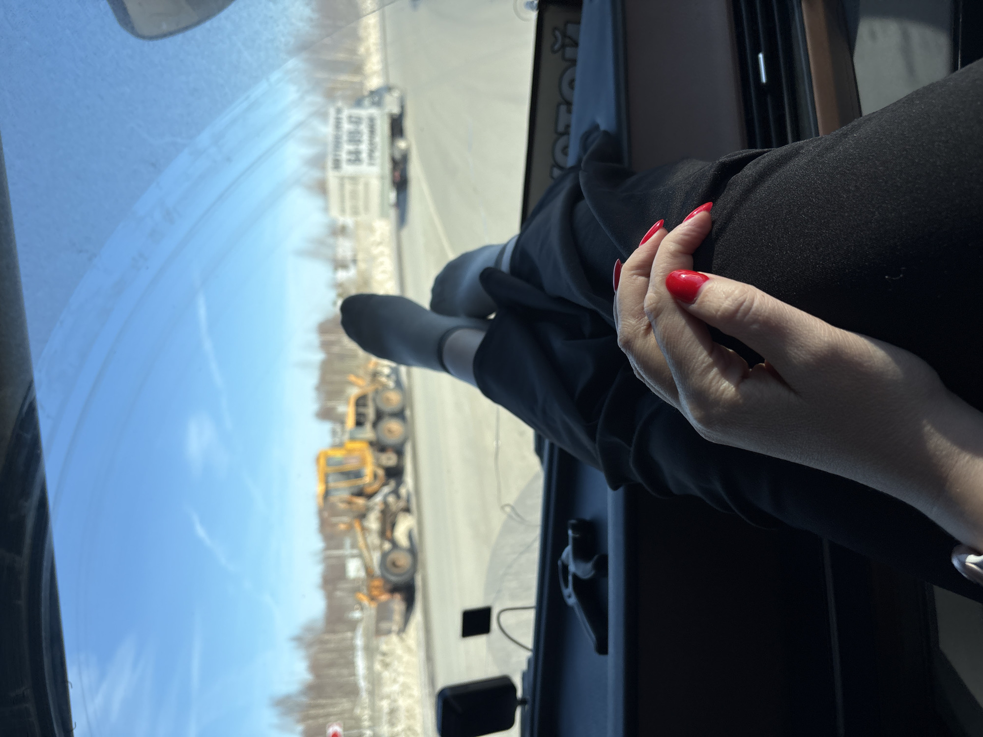 The image displays a scene viewed from the interior of a vehicle in Staroe Mikhailovskoe, Russia, looking forward through the windshield on a clear, sunny day.
In the foreground, a person's legs, clad in dark trousers and light gray socks, are crossed at the ankles and rest on the dark-colored dashboard. Below the legs, a right hand with bright red polished fingernails and a silver-toned ring on the ring finger is visible, resting on the person's lap. The vehicle's dashboard features a dark, textured surface, air vents, and a brown trim. Small black electronic devices or mounts are affixed to the dashboard near the windshield. Partially visible Cyrillic text, "ОТОЙ," is present on a label near the bottom of the windshield.
Outside the vehicle, a paved road is visible, flanked by residual snow on the shoulders. A large yellow road grader or construction vehicle is positioned on the right side of the road, appearing stationary. Further down the road, a white box truck, featuring the Cyrillic text "АВТОПЕРЕВОЗКИ" (Auto Transport) and the numbers "64-89-47" on its side, is also visible. Bare deciduous trees line the distant horizon under a bright blue sky with faint cirrus clouds. A red and white road sign is partially visible on the far left.