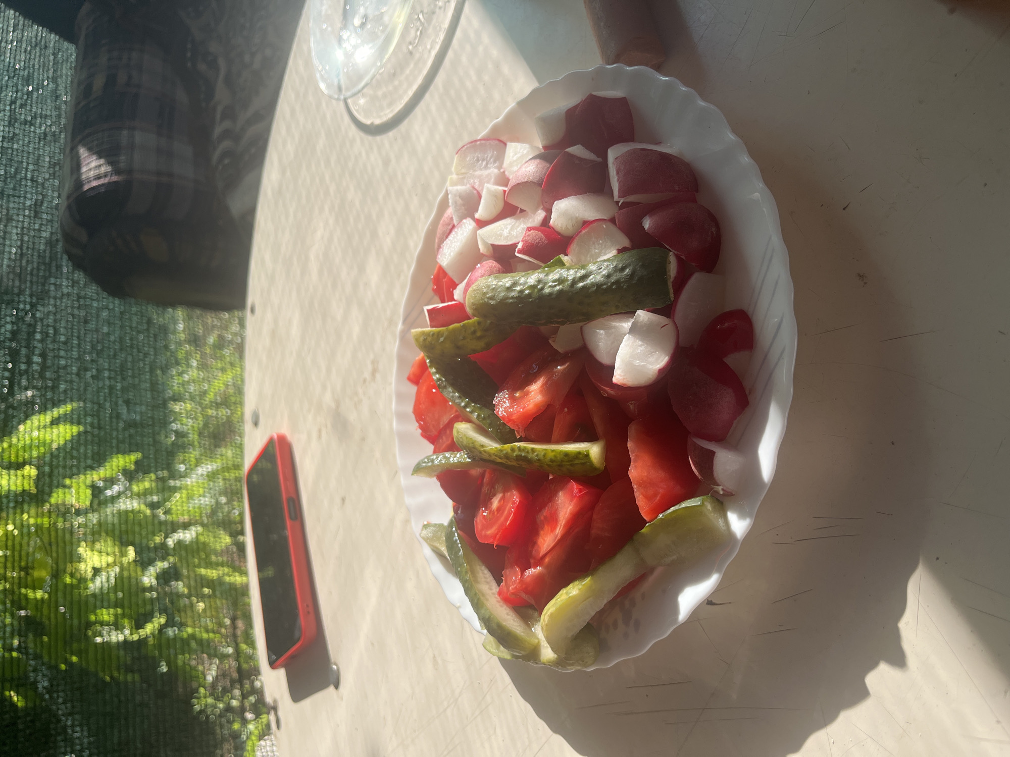 The image depicts a close-up of a white plate filled with a fresh salad consisting of chopped tomatoes, sliced cucumbers, and diced radishes. The salad is placed on a light-colored, possibly plastic, outdoor table. In the background, out-of-focus elements include green foliage, suggesting an outdoor setting with trees and possibly a privacy screen. A red smartphone is visible on the table to the left of the salad. The scene is bathed in sunlight, creating distinct shadows on the table surface. The location is specified as Rakhinka, Russia.