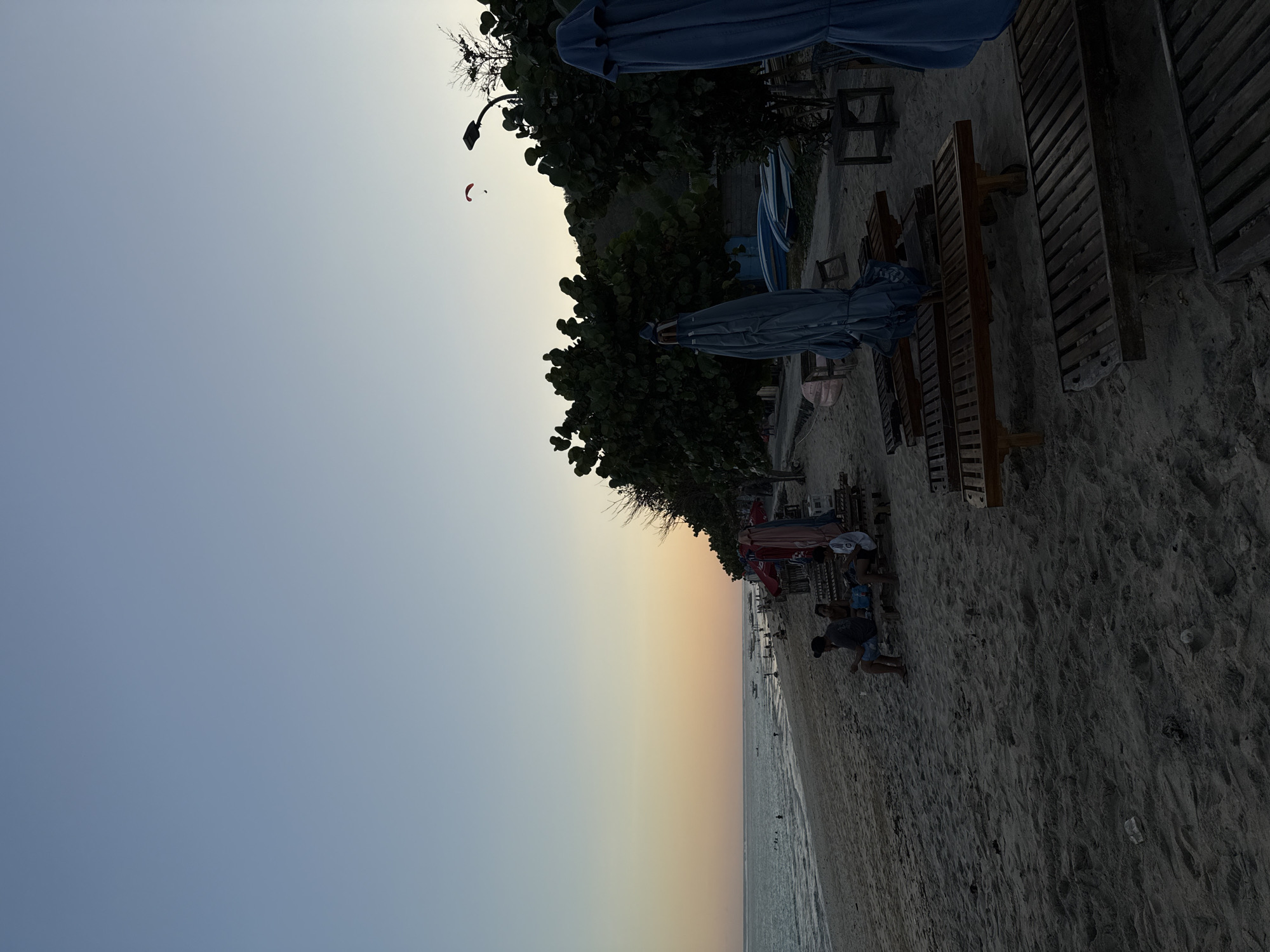 The image presents a wide view of a sandy beach at dusk in Kuta Selatan, Indonesia. The sky exhibits a gradient, transitioning from a light blue at the zenith to a warm orange-yellow near the horizon line. A solitary red paraglider with a dark figure beneath it is airborne against the sky in the upper right quadrant.
The foreground on the right side is dominated by multiple wooden beach loungers, some of which are partially obscured by large, closed blue beach umbrellas. Further back, along the treeline, more wooden loungers and benches are visible, with several individuals seated on them, engaged in passive activity. The sandy beach occupies the lower and mid-ground, showing faint footprints and scattered debris, including a plastic bottle.
Towards the left, the calm ocean extends to the horizon, reflecting the subdued light. Multiple individuals are visible swimming or wading in the nearshore waters, and several small boats are anchored further out to sea. The boundary between the beach and the land is marked by a line of dense, dark green trees, behind which a streetlamp with a single light fixture is visible on the upper right. On the far right, partially visible behind the trees and umbrellas, are several small boats or paddleboards stored on land, suggesting watersport activity. A gentle slope of land or hill is discernible on the right horizon, mostly silhouetted against the sky.