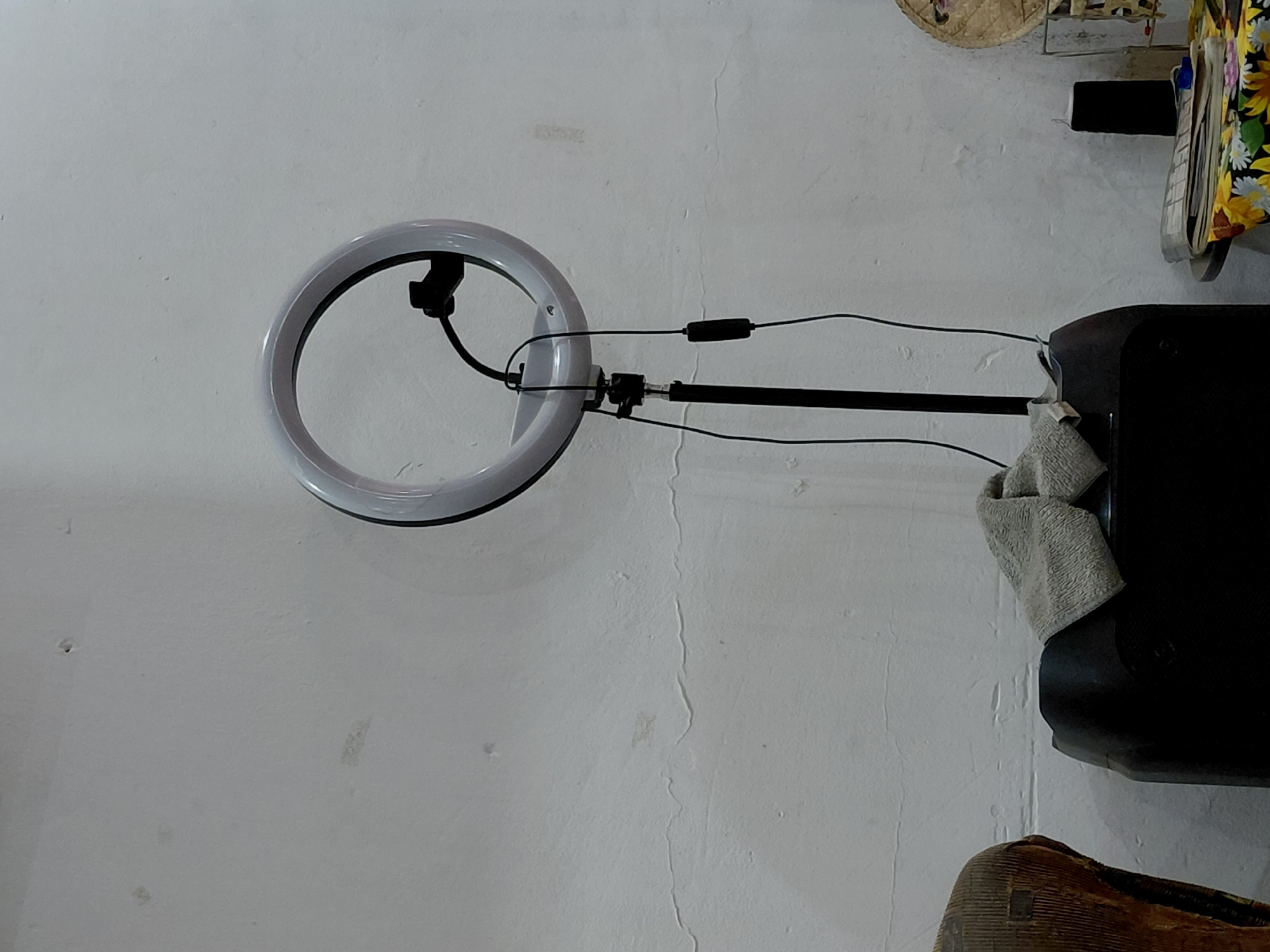 An indoor scene in Santa Ana, Venezuela features a ring light attached to a stand, positioned against a textured white wall. The ring light, with its circular form and internal components, is connected by cables that run along the stand. One cable includes an inline control module. The stand appears to be secured to a larger, dark, rectangular object, possibly a speaker or electronic device, over which a light-colored, folded cloth or towel is draped.
In the upper right corner of the frame, partially visible items include a woven, straw-like object (possibly a basket or hat), and a piece of fabric with a yellow and green floral pattern, beneath which white pages or books can be discerned. The bottom right corner shows a portion of a brown, textured object, likely a piece of furniture such as a chair. The white wall exhibits visible imperfections, including minor cracks and variations in its surface texture, suggesting an older or well-used interior. No people are present, and there is no visible text. The setup indicates an arrangement for photography, video recording, or streaming.