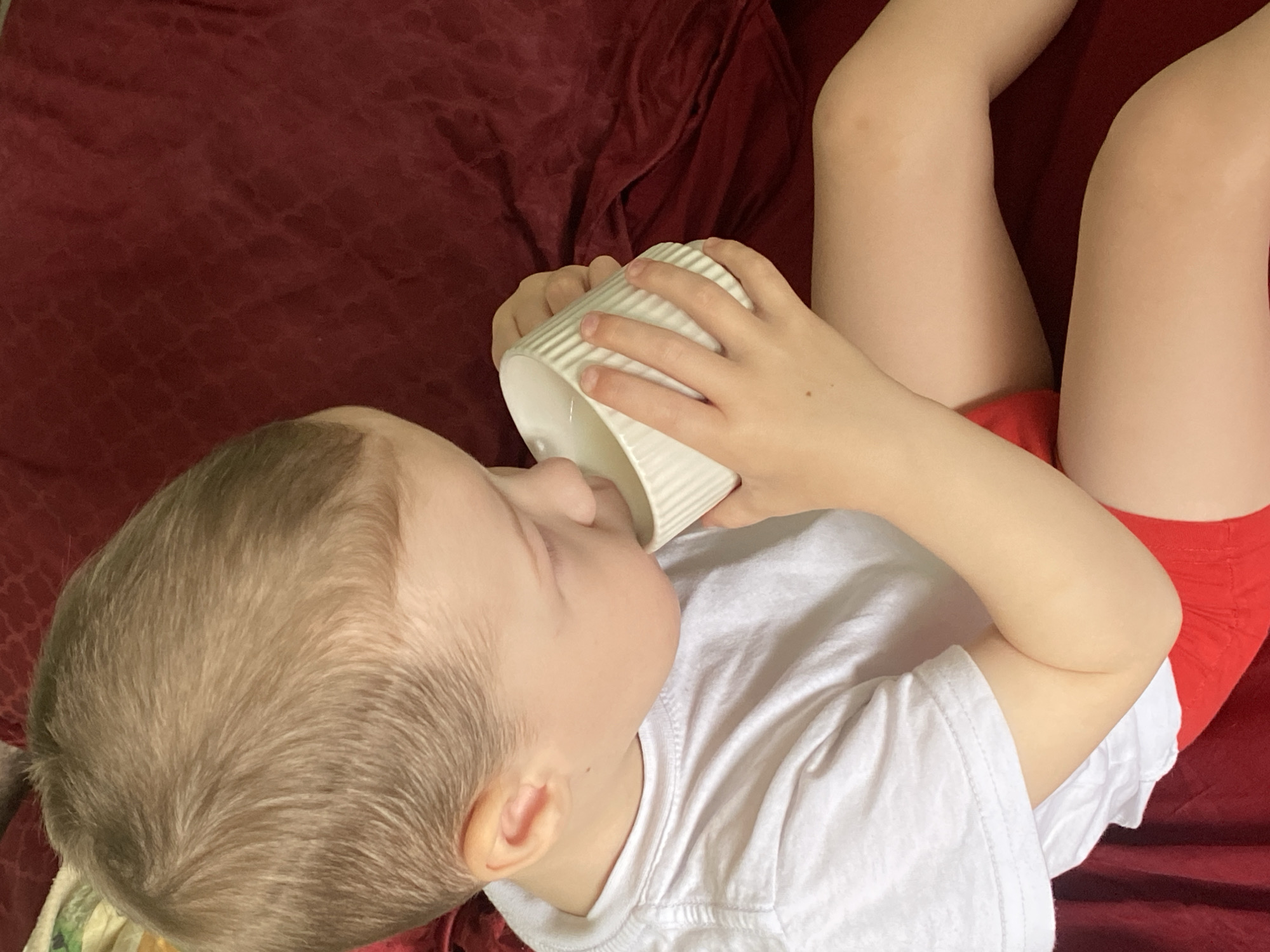 A young child is lying on a red fabric surface and drinking from a white mug. The child is wearing a white t-shirt and red shorts. The location is not discernible as Sochi, Russia.
