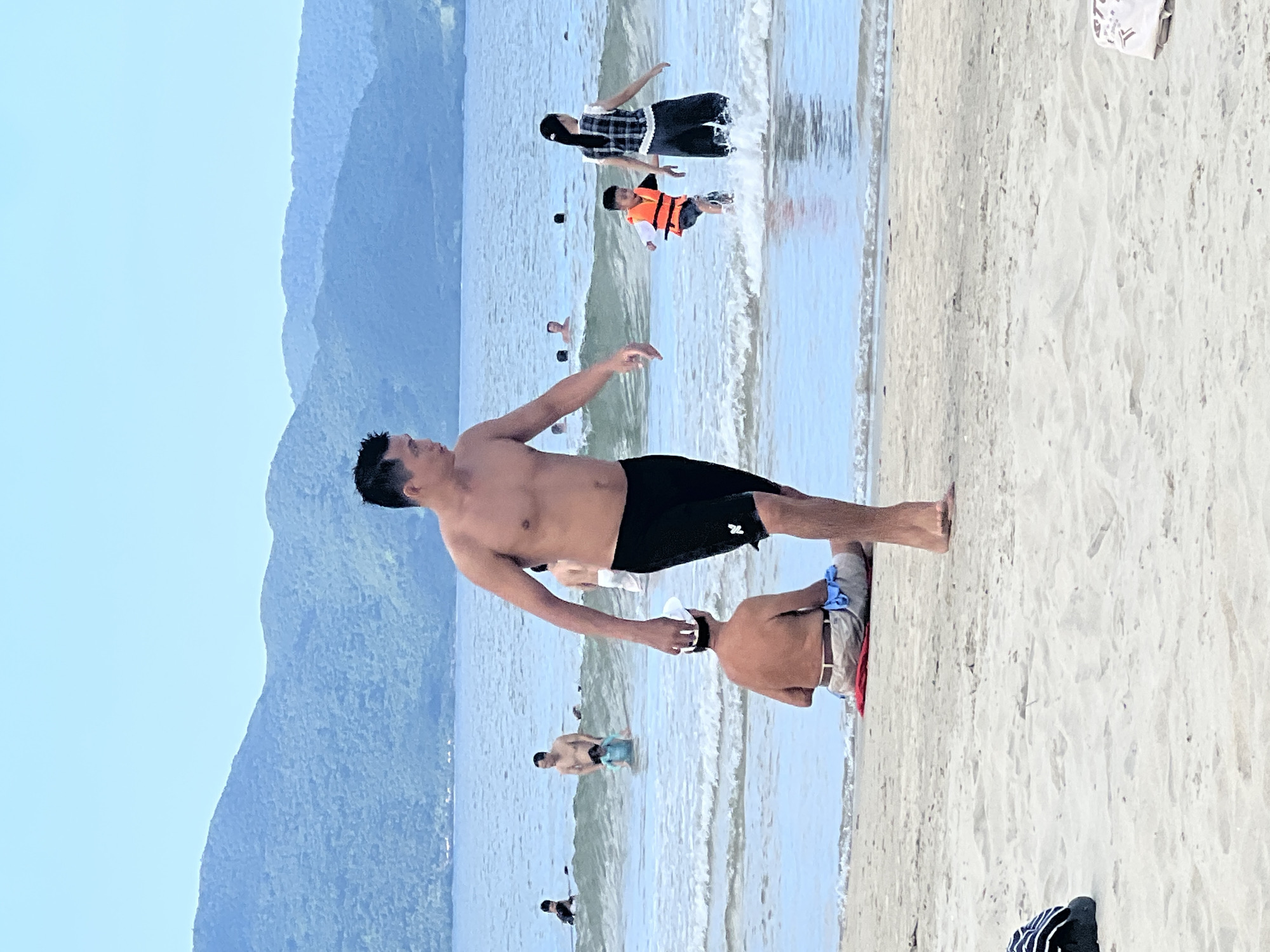 This image captures a vibrant scene at a beach in Đà Nẵng, Vietnam. In the foreground, a man with dark hair is walking on the sand, wearing black swim trunks. He is shirtless, and his body is tanned. To his left, a man is sitting on the sand, facing away from the camera, wearing shorts and a white hat. In the background, several people are in the shallow water. A child in an orange life jacket is running towards the shore, accompanied by a woman in a plaid top and dark pants. The water is calm with small waves. Behind the beach and water, lush green mountains form a scenic backdrop under a clear blue sky. The overall atmosphere is one of a pleasant summer day at the beach.
