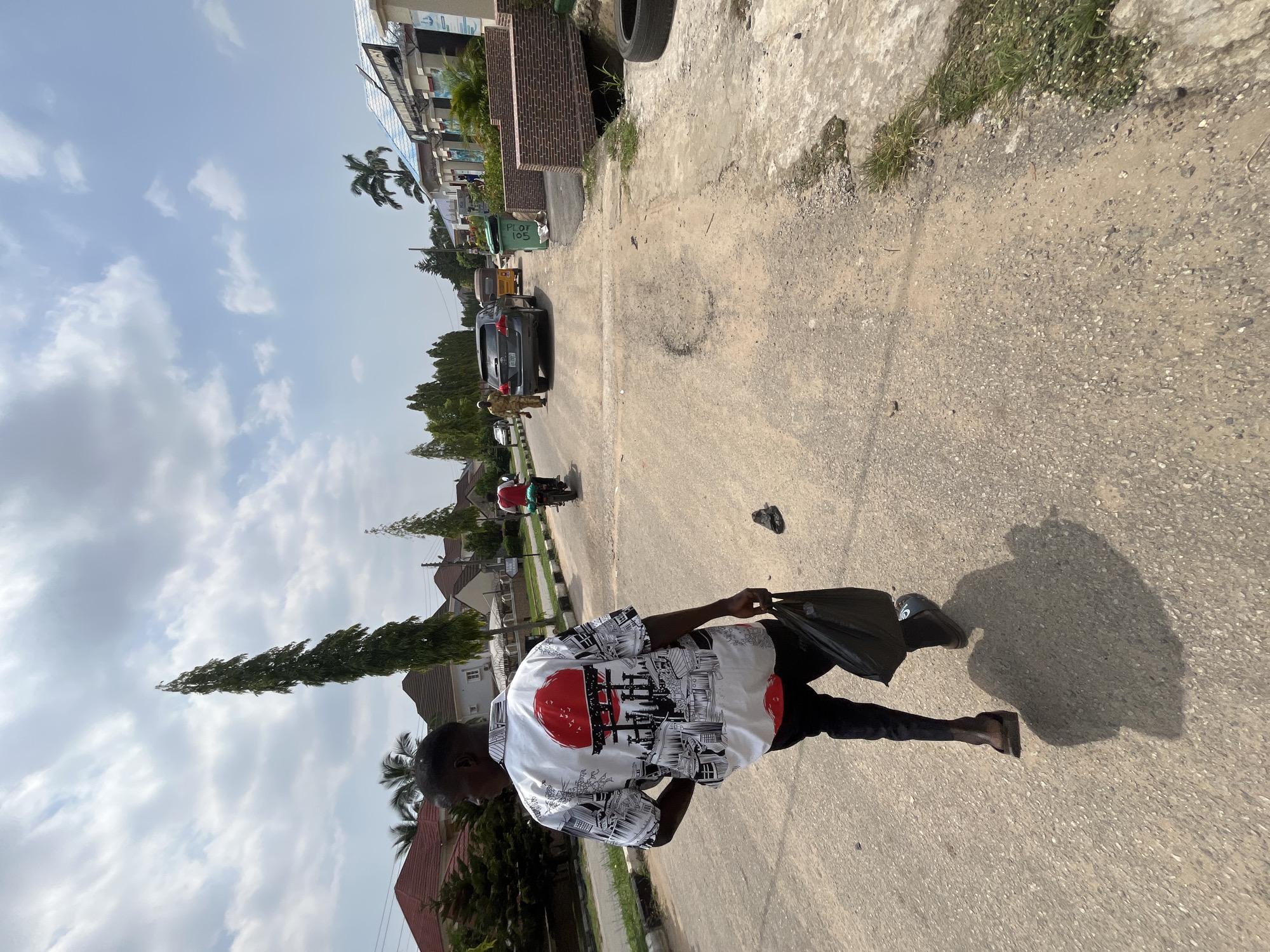 In Abuja, Nigeria, a person walks down a dusty, paved road. The individual, seen from behind, wears a white short-sleeved shirt with a red and black Japanese-inspired graphic, dark pants, and sandals. They are carrying a black plastic bag in their right hand. The street is lined with residential buildings and lush trees, including a tall, slender tree on the left and palm trees further down. Several vehicles are present, including a gray SUV, a tricycle, and motorcycles. The sky is partly cloudy with a bright sun. The road surface shows signs of wear and tear, with visible patches and cracks.
