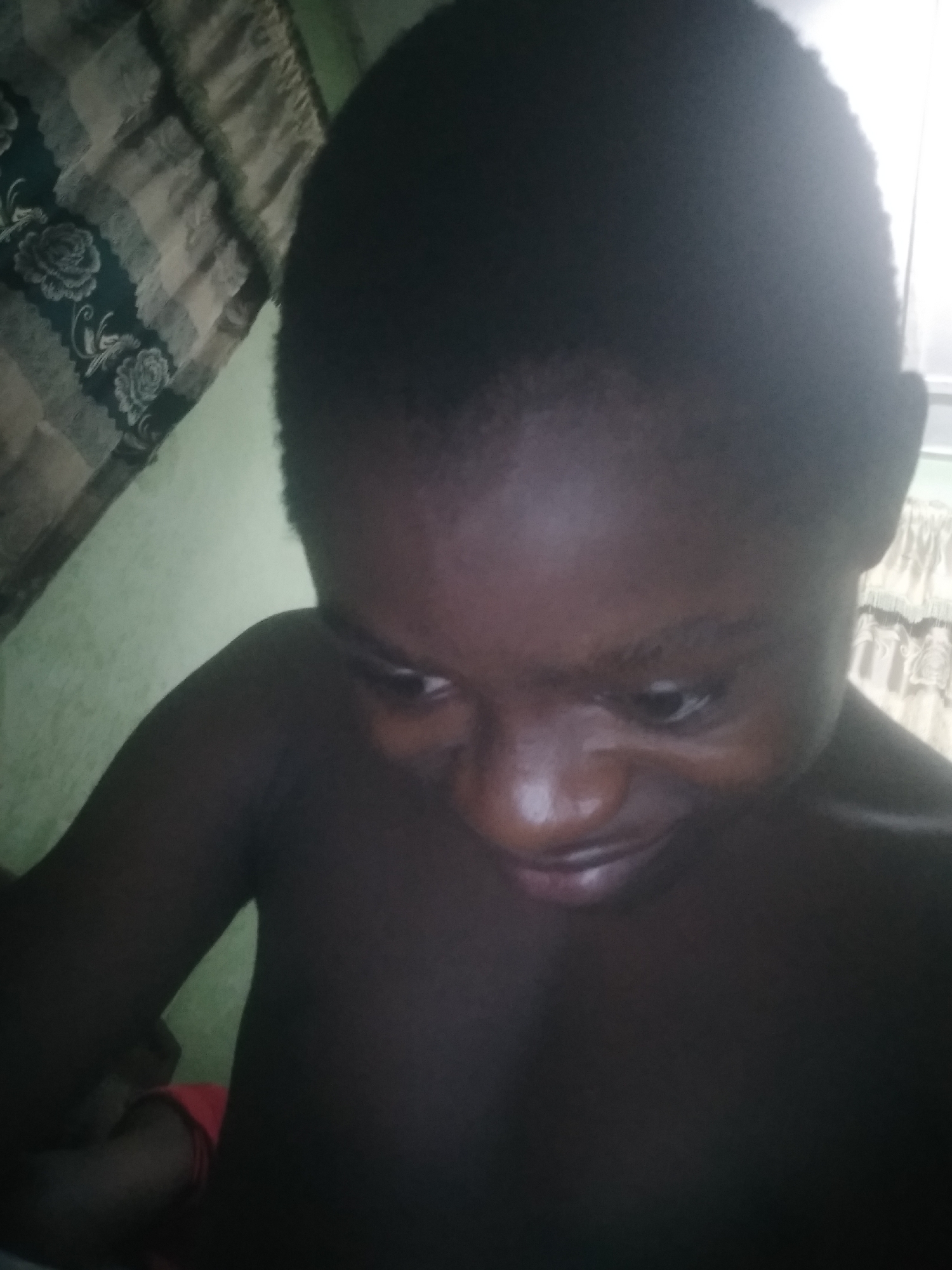 A medium close-up shot shows a person with dark skin looking down and slightly to the left. The person has short, dark hair. The environment appears to be indoors, as evidenced by a curtain with a floral pattern visible in the background. There is no clear evidence to verify the location as Shangisha, Nigeria.