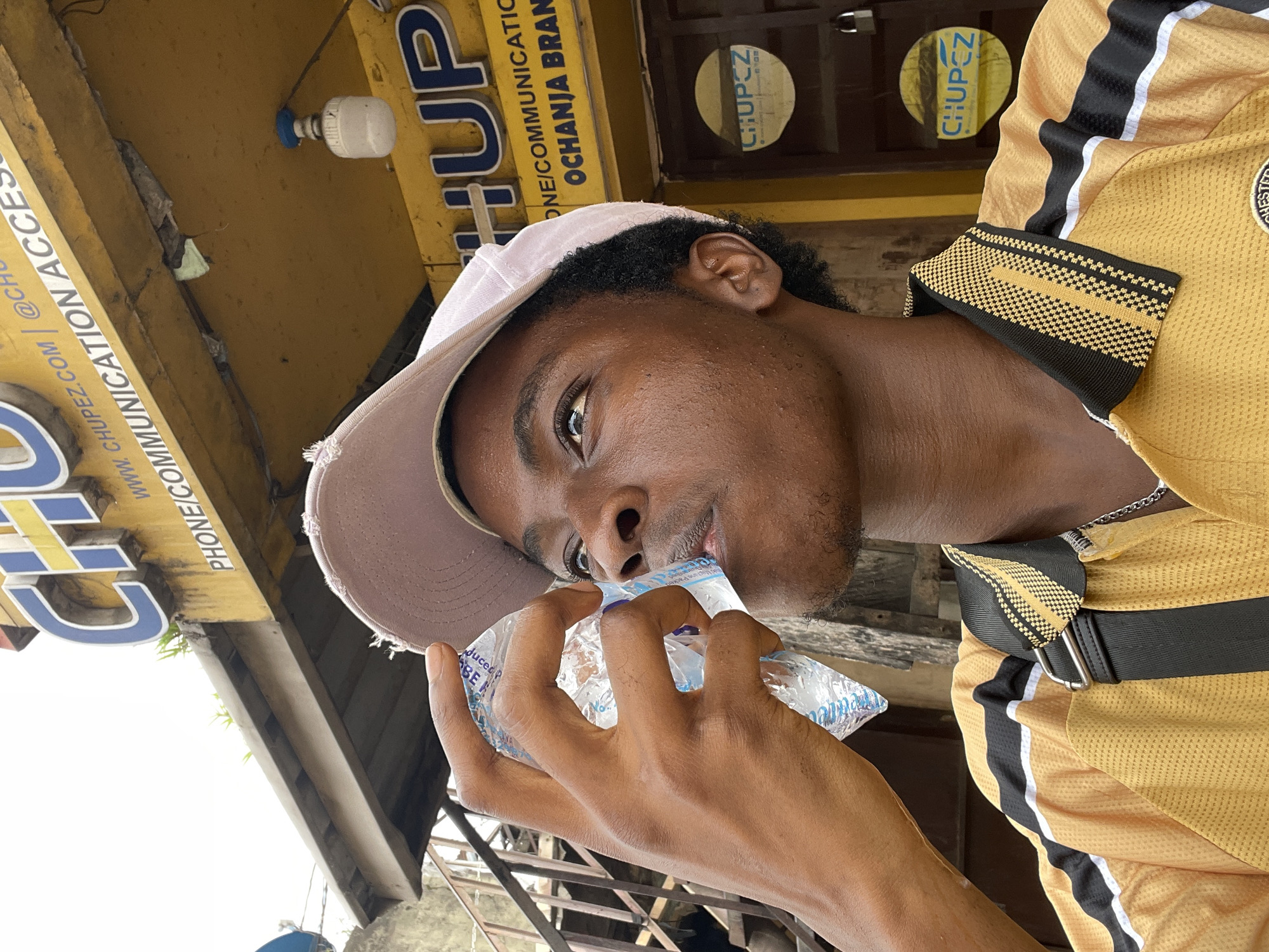 A young man is drinking water from a sachet. He is wearing a light pink baseball cap and a mustard-colored jersey with black and white accents. He appears to be standing outside of a business. The business sign says "CHUPEZ" in blue letters, along with the website, social media handle, and "PHONE/COMMUNICATION ACCESSORIES." The business location is listed as Ochanja branch. The location is possibly in the city of Fegge, Nigeria.