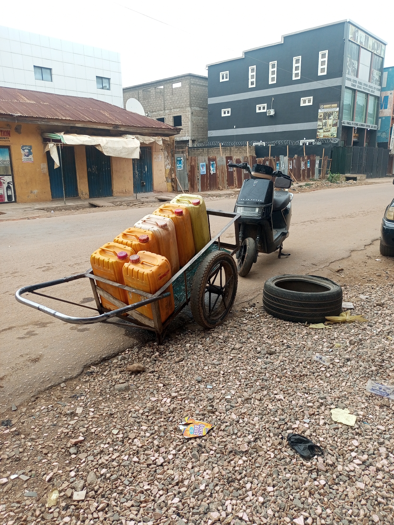 The image captures a street scene in Maiburiji, Nigeria. A black electric scooter is parked on the side of the road, hitched to a two-wheeled cart filled with about six yellow jerrycans. Next to the cart, a tire sits on a gravel area along the edge of the road. A black face mask lies on the gravel.
The background features buildings of varying sizes and conditions. A yellow building with a rusty metal roof is on the left side of the image. A multi-story grey building with white-framed windows is behind the scooter. Fences run along the edge of the street. The street itself is paved but appears worn.