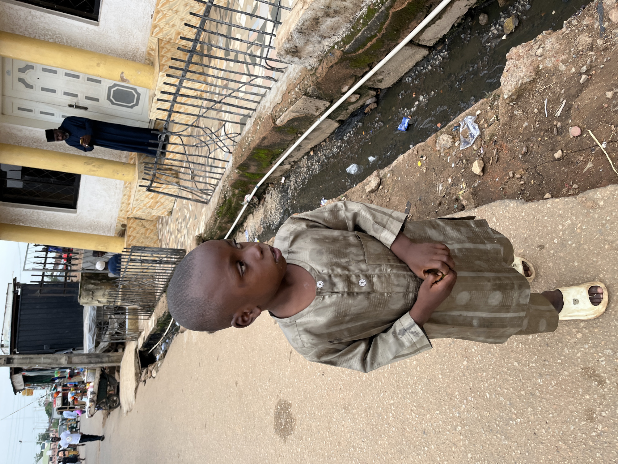 A young boy in a muted green traditional outfit stands on a dusty street. He wears simple, light-colored sandals. His gaze is directed to the right. The street appears to be a thoroughfare in an urban setting, with buildings lining one side. A drain runs alongside the street, filled with murky water and scattered debris. In the background, other people and a market stall are visible, suggesting a busy commercial area. Further down the street, a man in a dark robe and cap stands near a building entrance. The scene is set in Gura Top, Nigeria.