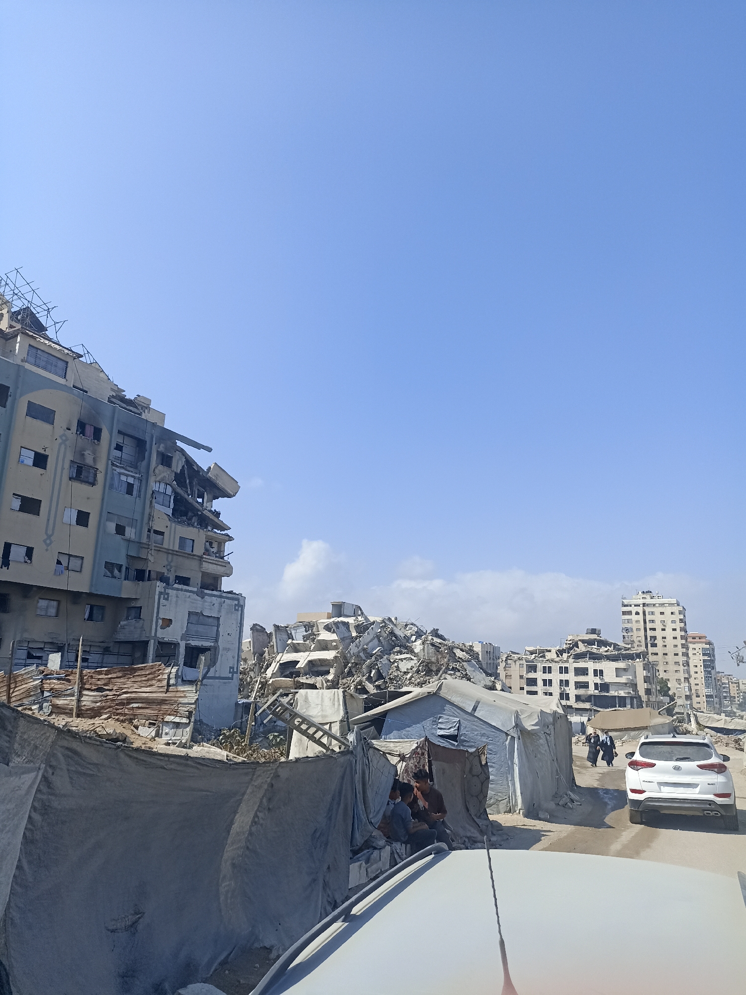 This image depicts a street scene in Gaza City, Palestinian Territories, characterized by significant destruction and makeshift shelters. In the foreground, the hood and windshield of a white vehicle are visible, suggesting the photo was taken from inside a car. To the left, a large, partially collapsed multi-story building exhibits severe damage, with exposed rebar and missing sections of its facade. Scaffolding is still attached to parts of this structure.
Further down the street, a large pile of rubble from destroyed buildings dominates the midground. Adjacent to the rubble are several tents and temporary structures made of tarpaulin, likely serving as shelters for displaced individuals. Some people are visible within these makeshift dwellings.
In the background, other damaged buildings and structures are present, indicating widespread destruction in the area. A white SUV is driving away from the camera on the right side of the street, and two figures, appearing to be women in traditional attire, are walking in the same direction. The sky is clear and blue. The overall scene conveys a sense of devastation and the immediate aftermath of conflict.