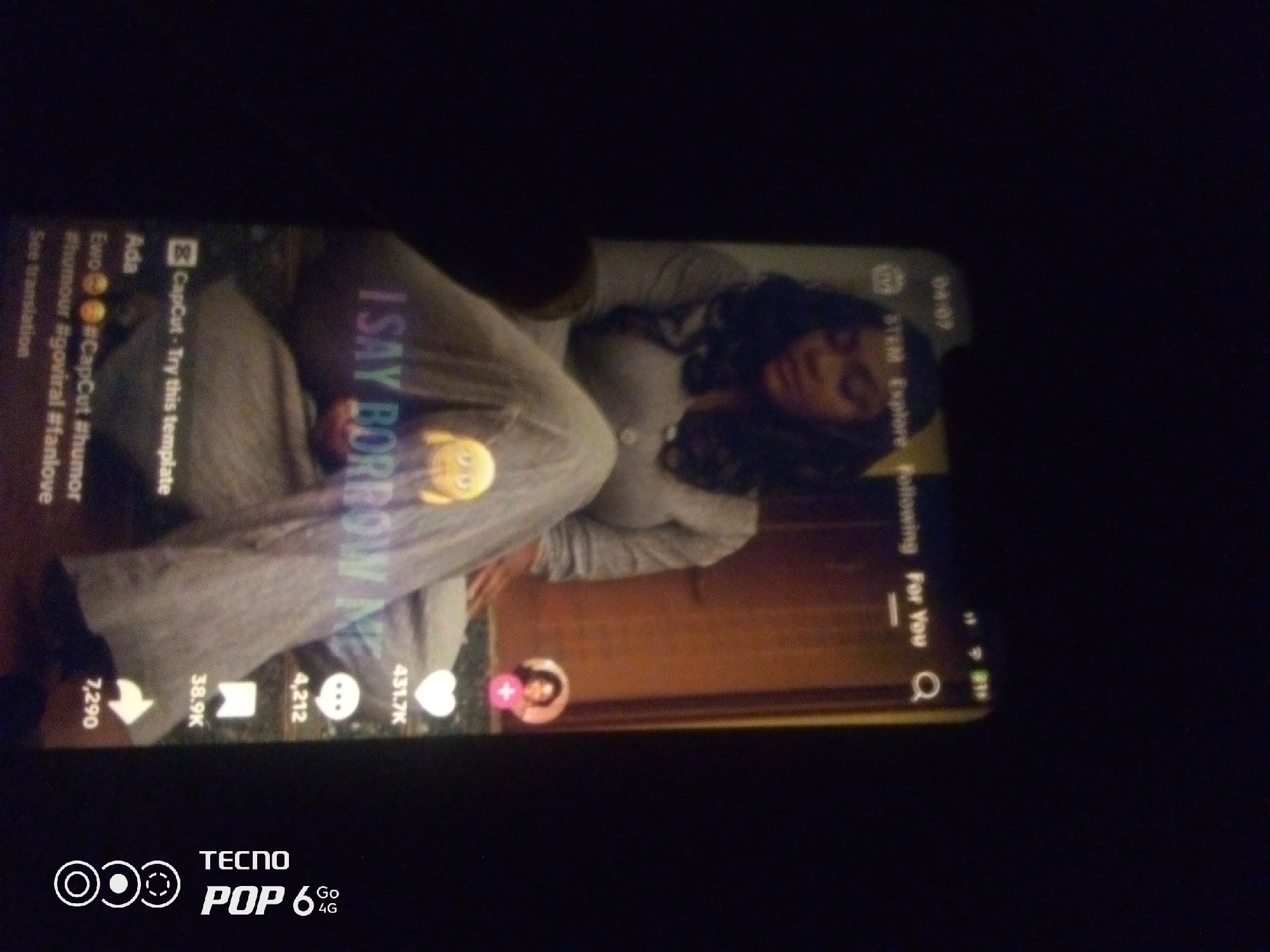 A person is lying on their side, wearing gray sweatpants and a gray hooded sweatshirt. They are looking at a phone screen, which displays a video with text overlay. The text on the screen says "I SAY BORROW MY HEART". The phone is held by the person, and the screen is illuminated. The background is dark and appears to be indoors. The bottom of the image shows the "TECNO POP 6 Go 4G" branding. There are no identifiable landmarks or contextual clues to suggest a specific location within Howayi, Nigeria. The image is a close-up shot of the person and the phone.