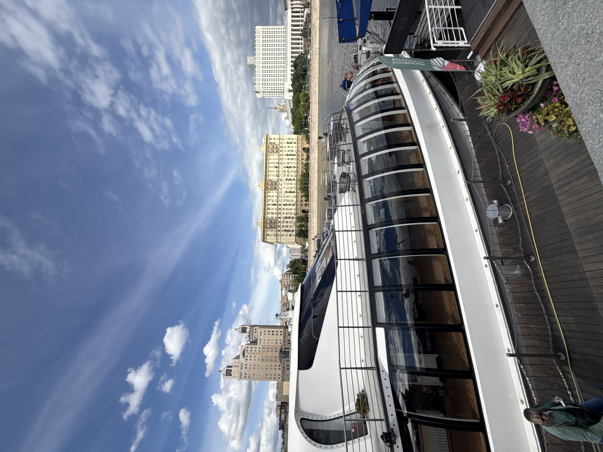 The image depicts a side view of a modern boat docked on a wooden platform next to a river. The boat features a curved, glass-enclosed structure. The sky is blue with scattered clouds. In the background are several tall buildings, including the White House in Moscow, Russia. Some greenery is visible between the buildings. On the platform, there are flowers in pots and a metal railing. A sign is visible, with text that includes "Московское". A person is partly visible in the bottom right corner.