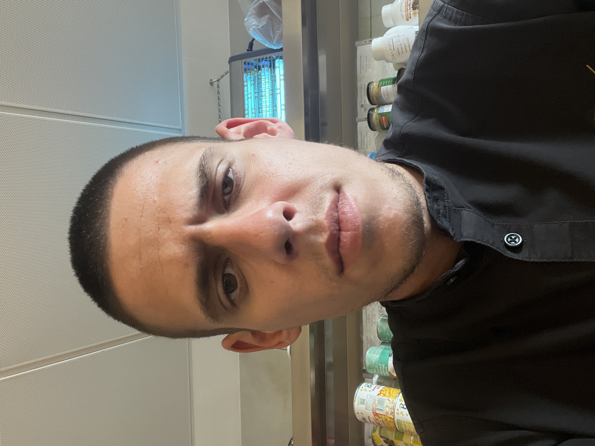 A close-up selfie of a man with short, dark hair and light skin. He is wearing a black collared shirt with a distinctive button. The background is blurry but appears to be an indoor commercial setting. There is a blue bug zapper hanging on a chain in the background, along with shelves containing various canned goods and bottled products. The wall behind the man is a white, perforated paneling. The location is not discernable based on the image, and information about Sofia, Bulgaria is not possible.