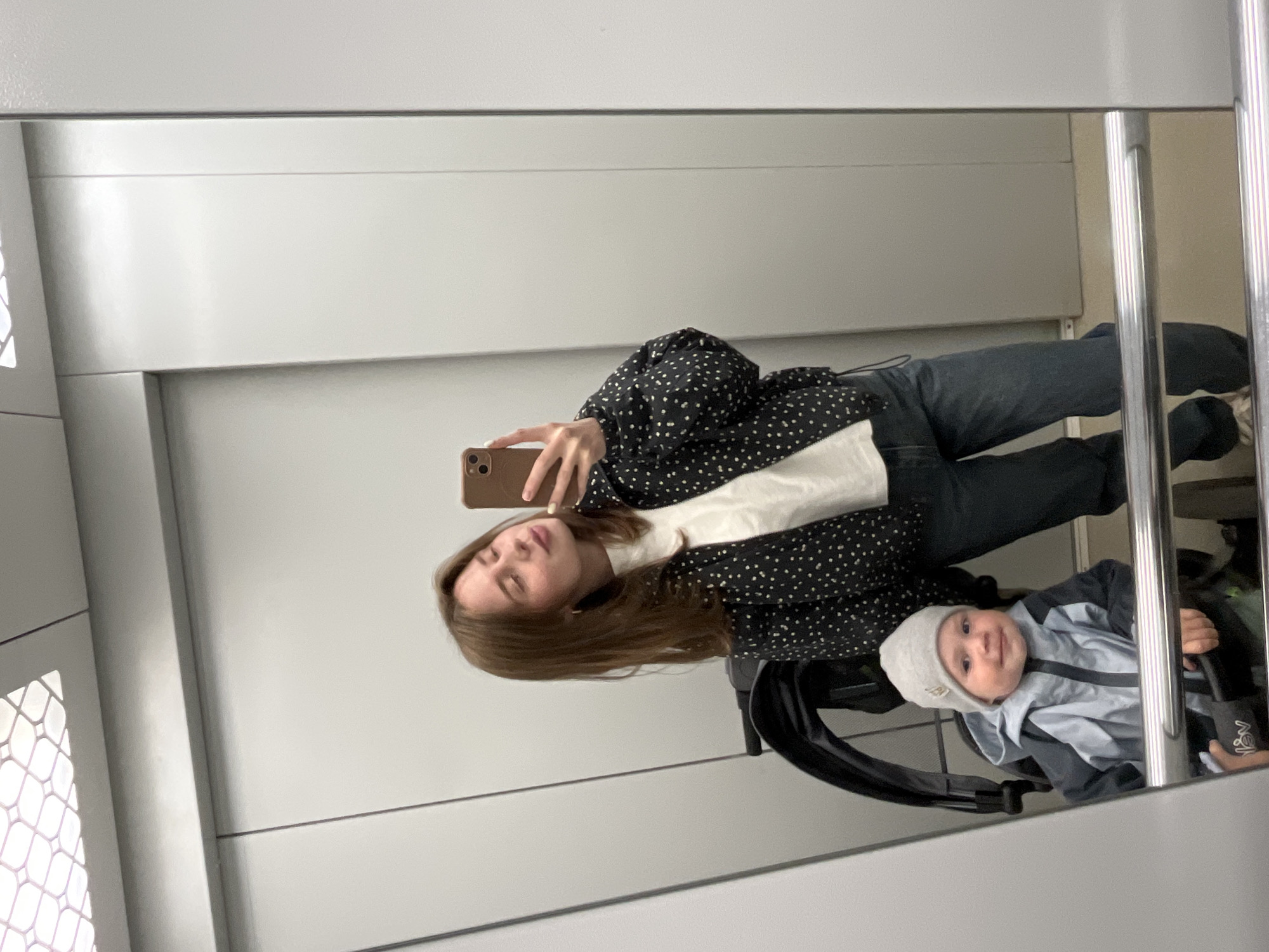 The image is a mirror selfie featuring a woman and a baby in a stroller, likely taken within an elevator or a public indoor space with reflective walls. The woman, with long brown hair falling to her left, holds a brown smartphone to capture her reflection. She is wearing a black polka-dotted blouse over a white long-sleeved shirt and dark jeans. Her eyes are partially closed, conveying a calm or thoughtful expression.
Below and slightly to the right of the woman, reflected in the mirror, is a baby seated in a black stroller. The baby is dressed in a light blue and dark blue jacket and a light gray beanie, looking directly towards the camera with a gentle smile.
The setting consists of smooth, light gray or white paneled walls. On the left side of the reflection, a white decorative grate or vent with a geometric pattern is visible. On the far right, a vertical silver metal handrail or support structure is prominent, characteristic of an elevator interior or a public passageway. The overall lighting is even and appears artificial. There are no direct indications of the time of day or outdoor weather. While a small, dark logo might be present on the baby's beanie and a tag on the stroller, no text is clearly legible. The photo's context is Chaikovsky, Russia.