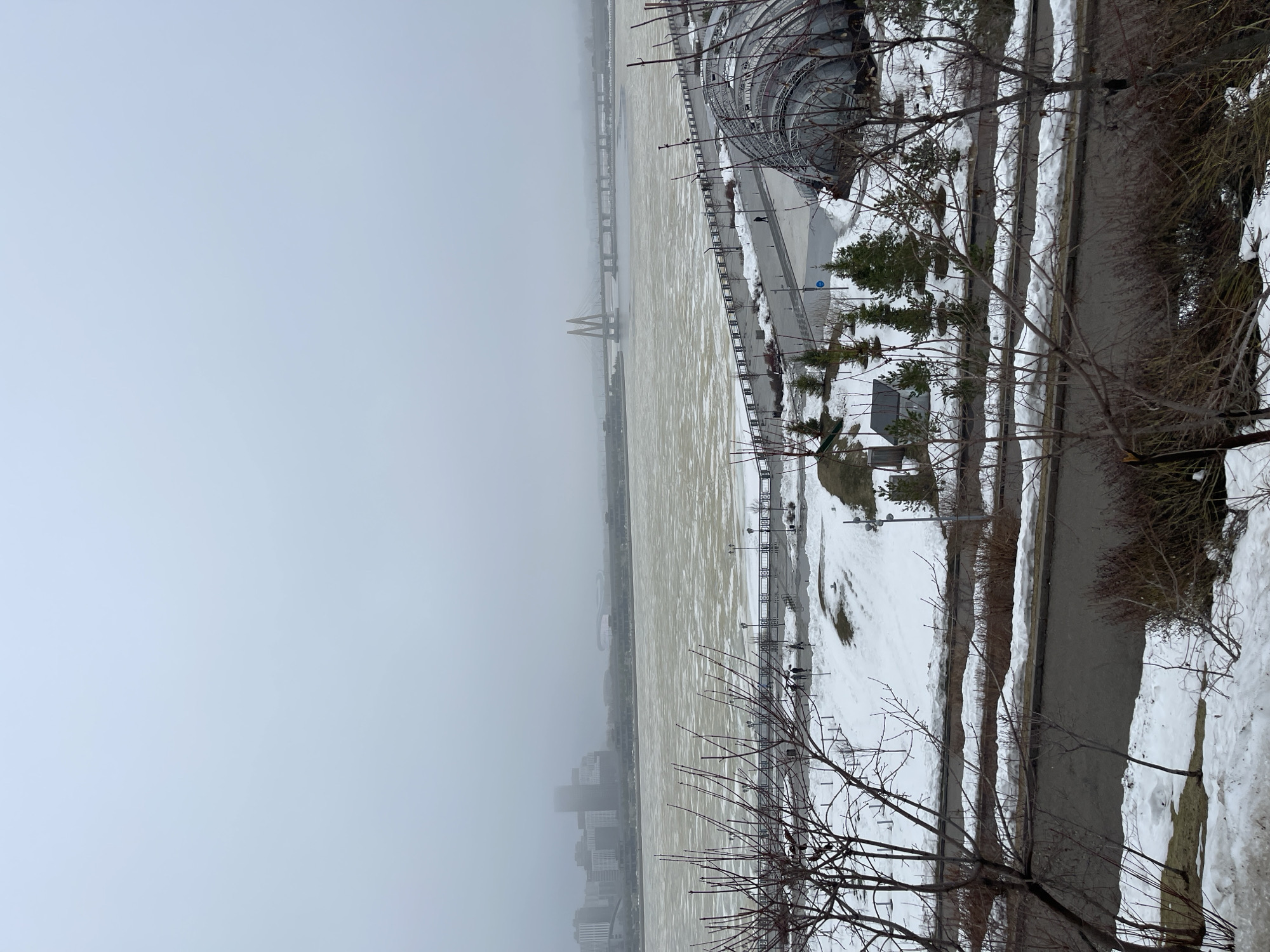 The image shows a winter scene in Kazan, Russia, looking towards the Kazanka River and the city skyline shrouded in fog or haze. The river is partially frozen, with a yellowish-brown, ice-covered surface. The Millennium Bridge is visible in the distance, spanning the river.
Along the riverbank is a landscaped area with patches of snow, bare trees, and some evergreen shrubs. A walkway runs parallel to the river, with structures that might be part of a park or recreational area visible. Some architectural structures are also visible, suggesting a park near the river.
The foreground features bare tree branches.