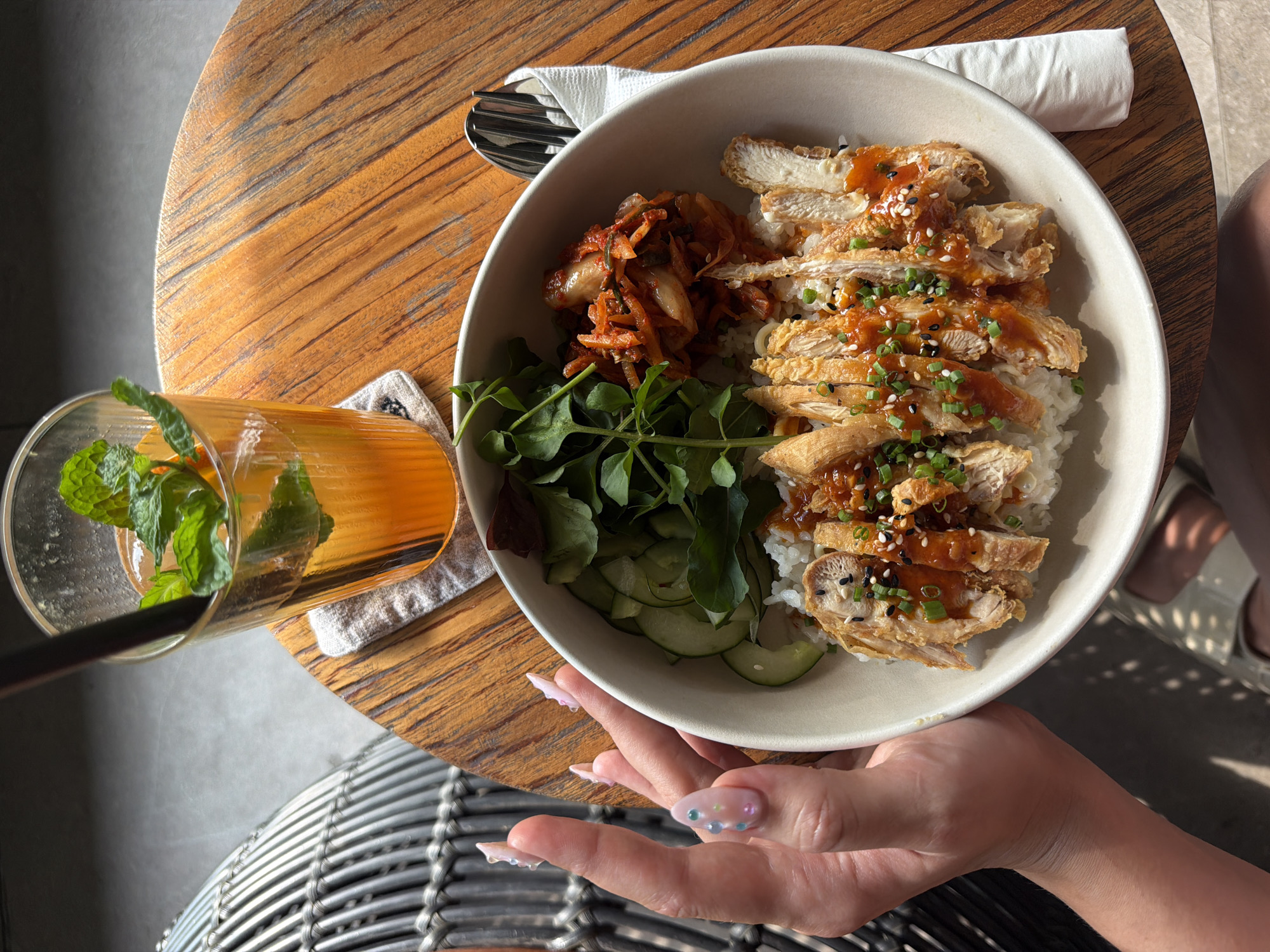 A person's hands are visible on the left side of the frame, with manicured nails adorned with small, iridescent beads. They are reaching towards a bowl of food on a wooden table. The bowl contains white rice topped with sliced, breaded chicken, drizzled with a reddish sauce and sprinkled with sesame seeds and green onions. The dish is accompanied by sliced cucumbers, a pile of kimchi, and a green leafy vegetable. To the upper right of the bowl is a tall glass filled with an orange beverage, ice, and fresh mint leaves, with a dark straw. The table is made of wood with a visible grain pattern and is illuminated by dappled sunlight. In the background, a woven chair and a hint of a building's interior are discernible. This scene is located in North Kuta, Indonesia.