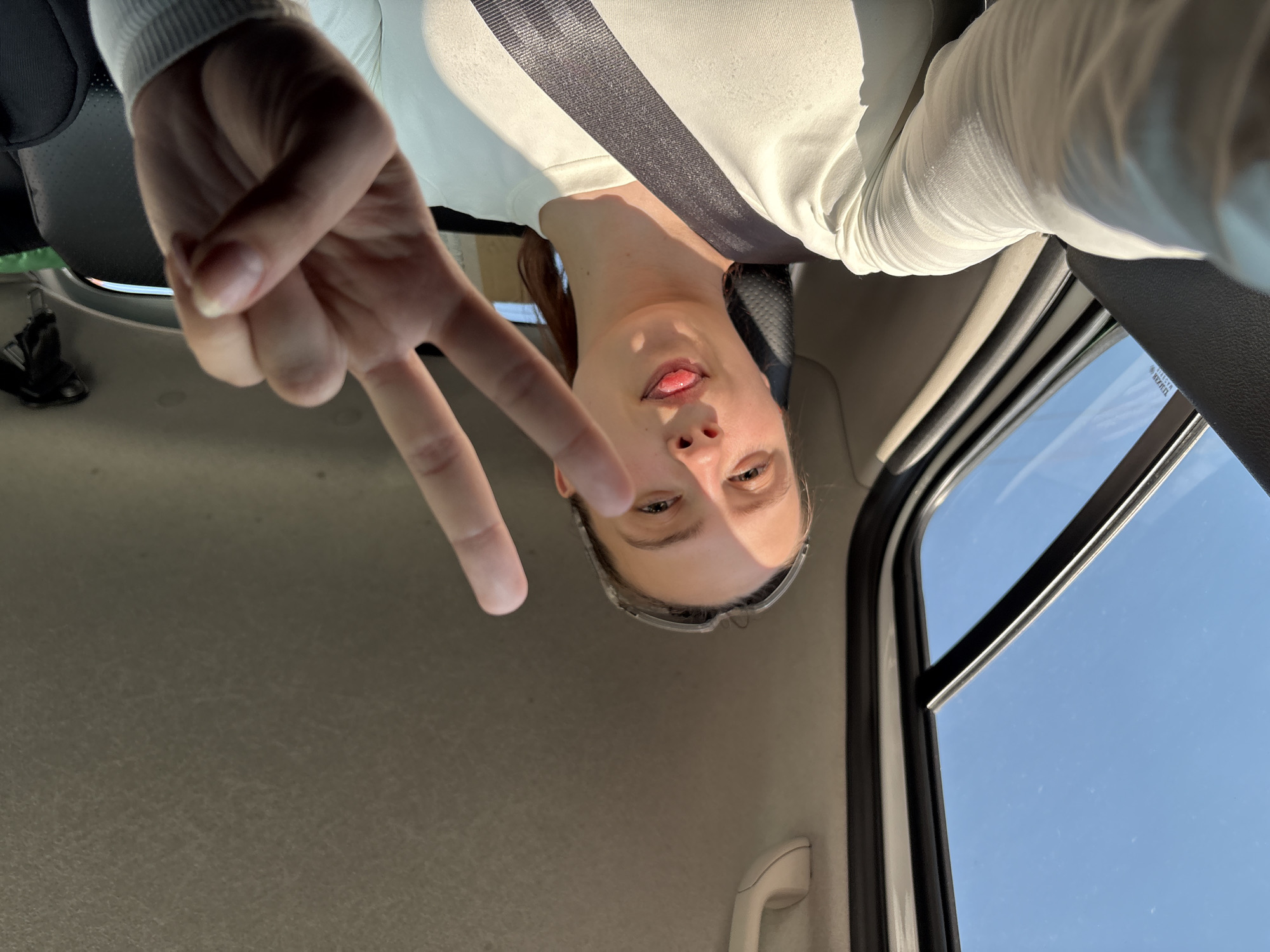 A woman is sitting in the driver's seat of a car, wearing a white long-sleeved shirt and a seatbelt. She is holding up a peace sign with her left hand. The photo is taken from a low angle, looking up at the woman. The car window shows a clear blue sky. The interior of the car is beige. The location cannot be determined from the image.
