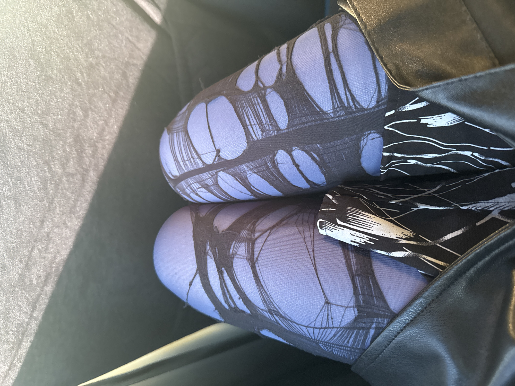 A close-up photograph shows two legs covered in blue and black tights that are ripped or designed to appear ripped. A black leather garment, potentially a coat or jacket, is positioned beside the legs. There is no indication the photograph was taken in Krasnoyarsk, Russia.