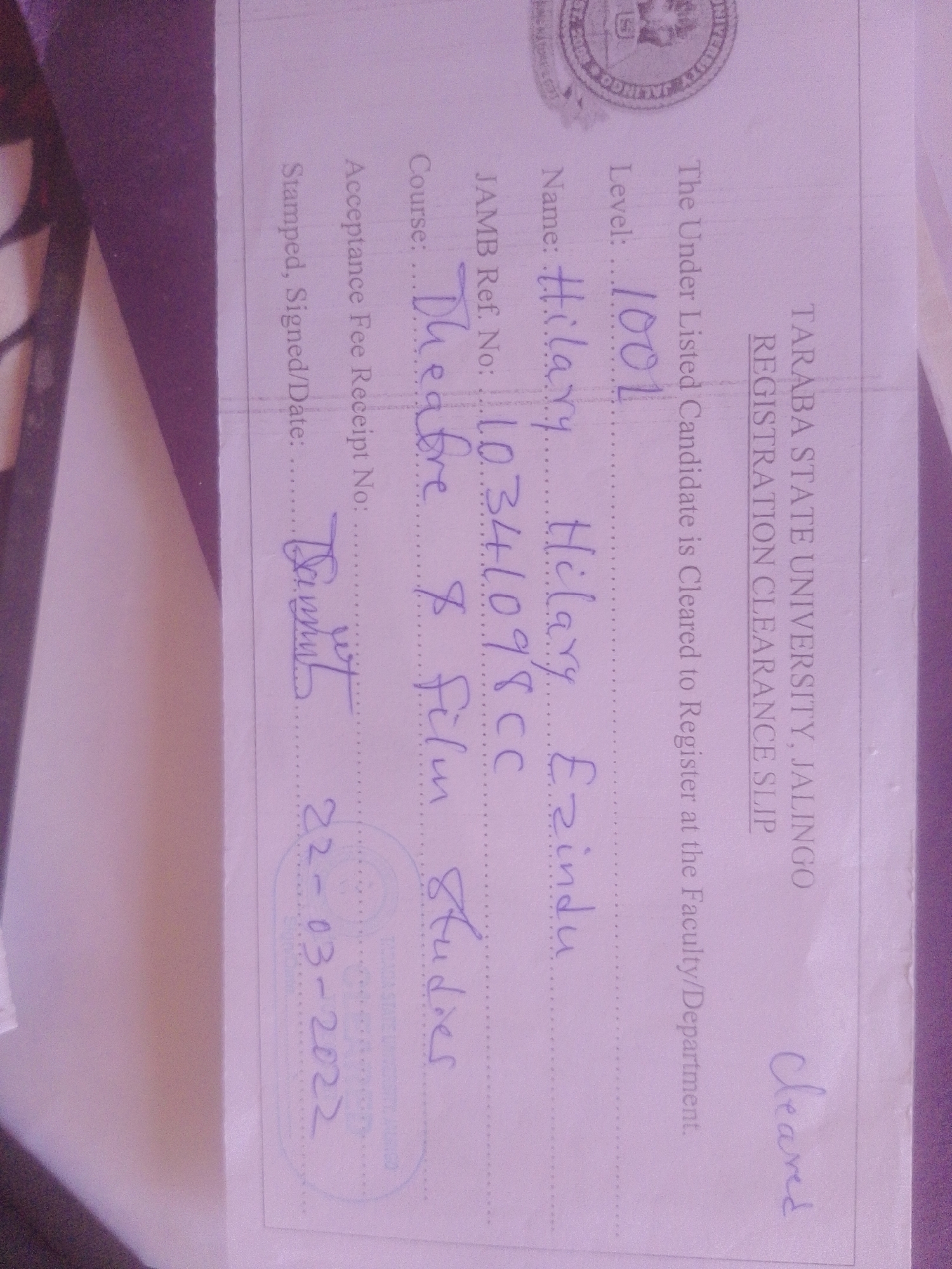 The image is a scanned document, likely an official clearance slip from Taraba State University, Jalingo, Nigeria. The document is printed with lines and headings. Handwritten text includes a candidate's name, Hilary Ezindu, a JAMB reference number (10344098CC), a course title (Theatre & Film Studies), and a level (100L). There is also a handwritten date, 22-03-2022, and a signature that appears to be "Dammit". The document is stamped with what appears to be the university's seal. The background shows part of a dark, textured surface, possibly a folder or binder.