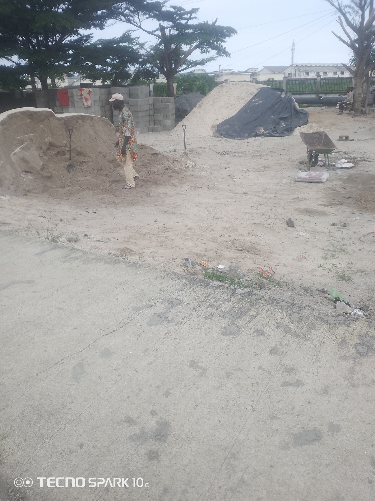 In Adesanya, Nigeria, a construction site is depicted under an overcast sky. The scene is dominated by piles of sand, a common material in construction. A man, dressed in a patterned shirt and light-colored trousers, stands near a large sand dune, seemingly involved in the construction activities. His attire suggests a practical choice for the environment.
Other objects present include stacks of concrete blocks, indicating the building materials readily available on site. A wheelbarrow, a shovel, and a large black tarp covering some materials are also visible, all essential tools and elements of a construction zone. In the background, trees provide a natural backdrop, with some residential buildings and power lines suggesting the site is in a populated area.
The overall impression is one of ongoing work, with the scattered debris and materials hinting at the dynamic nature of a construction project. The time of day appears to be daytime, based on the ambient light, though the cloudy weather obscures direct sunlight. The scene is characteristic of many developing areas where construction is actively underway. No specific emotional cues are evident from the subjects. The text "TECNO SPARK 10c" is visible at the bottom left corner of the image.