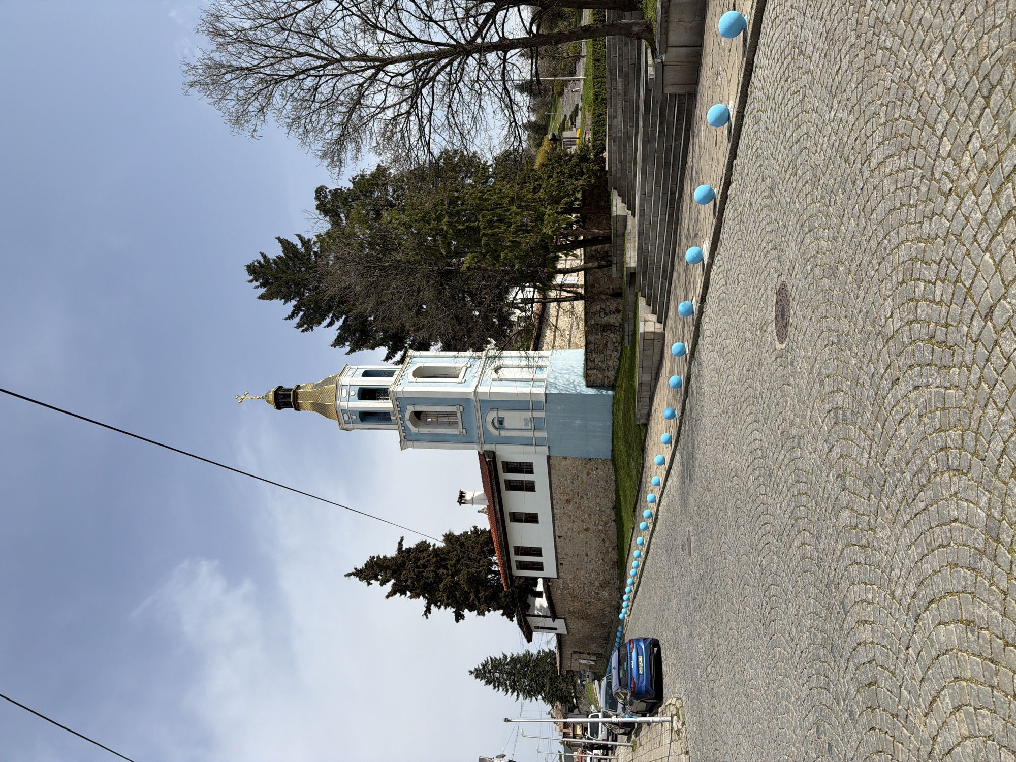 The image depicts the Assumption of the Virgin Mary Church in Panagyurishte, Bulgaria. The church's bell tower is visible, featuring a golden dome topped with a cross. The tower has blue and white accents, while the main building is white with a reddish-brown roof. The church is bordered by a stone wall and green vegetation. In the foreground, a cobblestone street is lined with evenly spaced, light-blue spheres. Cars are parked along the street. Above the church, tree branches reach into a slightly cloudy sky. A power line runs across the upper portion of the image.