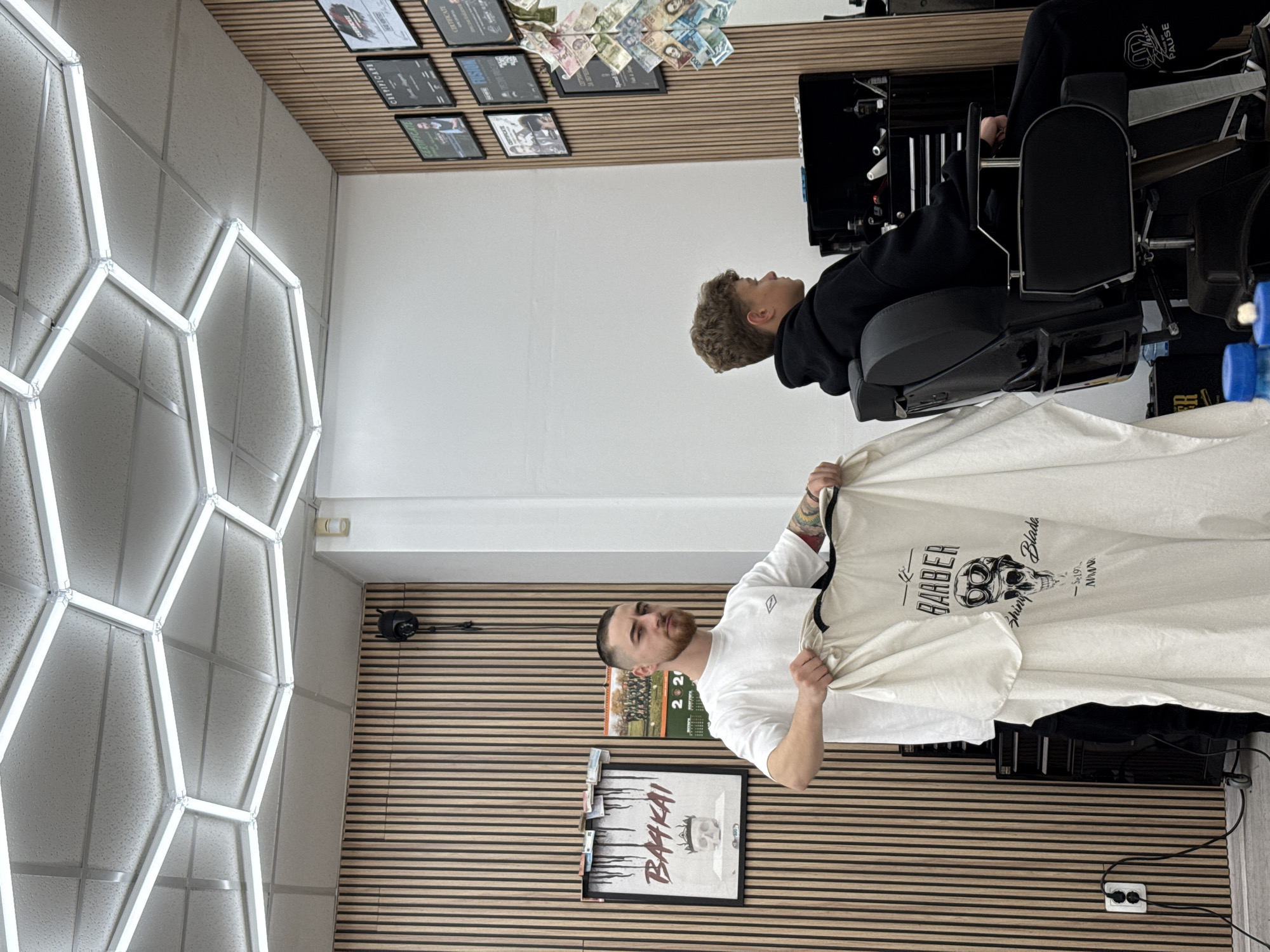 A barber shop in Sofia, Bulgaria. A male barber in a white t-shirt holds up a white barber cape with a skull graphic and "BARBER Shiny Blade" printed on it. A young male client sits in a black barber chair. The walls are decorated with framed posters and cash. The ceiling has a hexagonal lighting fixture.